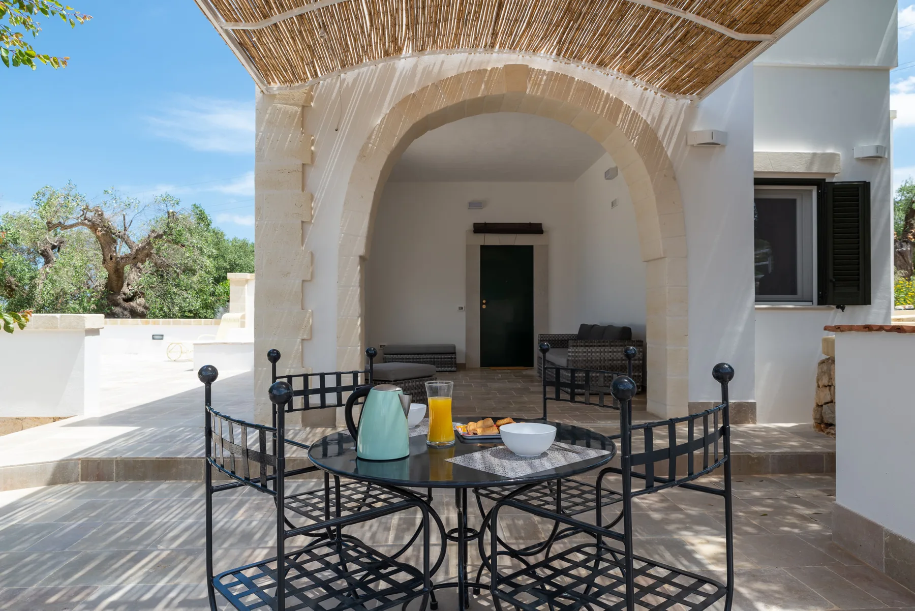 A shaded outdoor patio has a round table and black metal seating beneath an arched porch.
