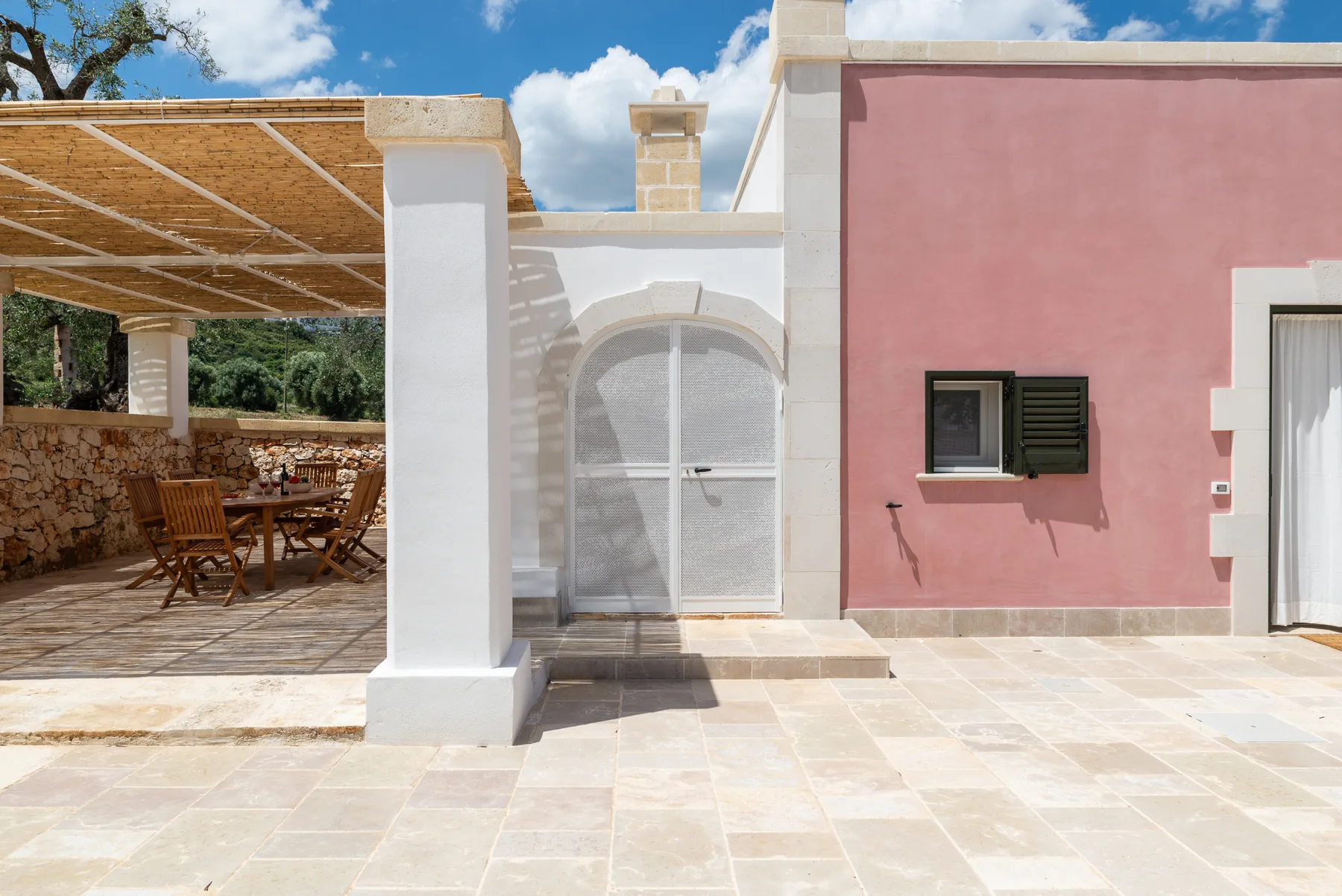 A sunlit stone patio beside a pink building, with shaded outdoor seating.