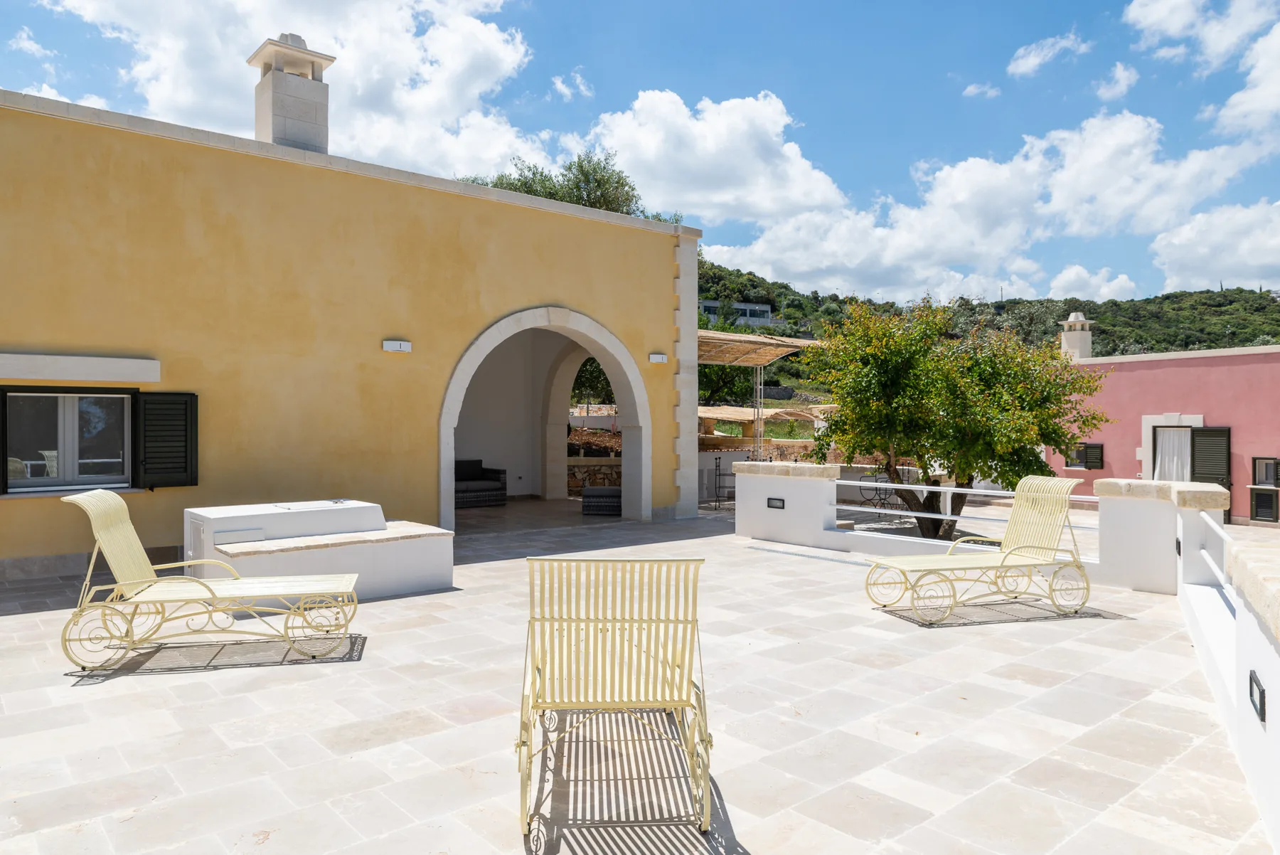A sunlit terrace with pale paving, loungers, and an arched building entrance.
