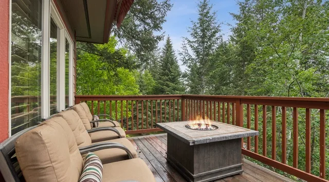 An outdoor deck has cushioned seating and a square fire table overlooking trees.