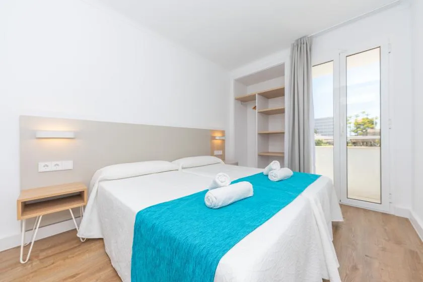A bright bedroom with white bedding and a turquoise bedspread beside tall windows.