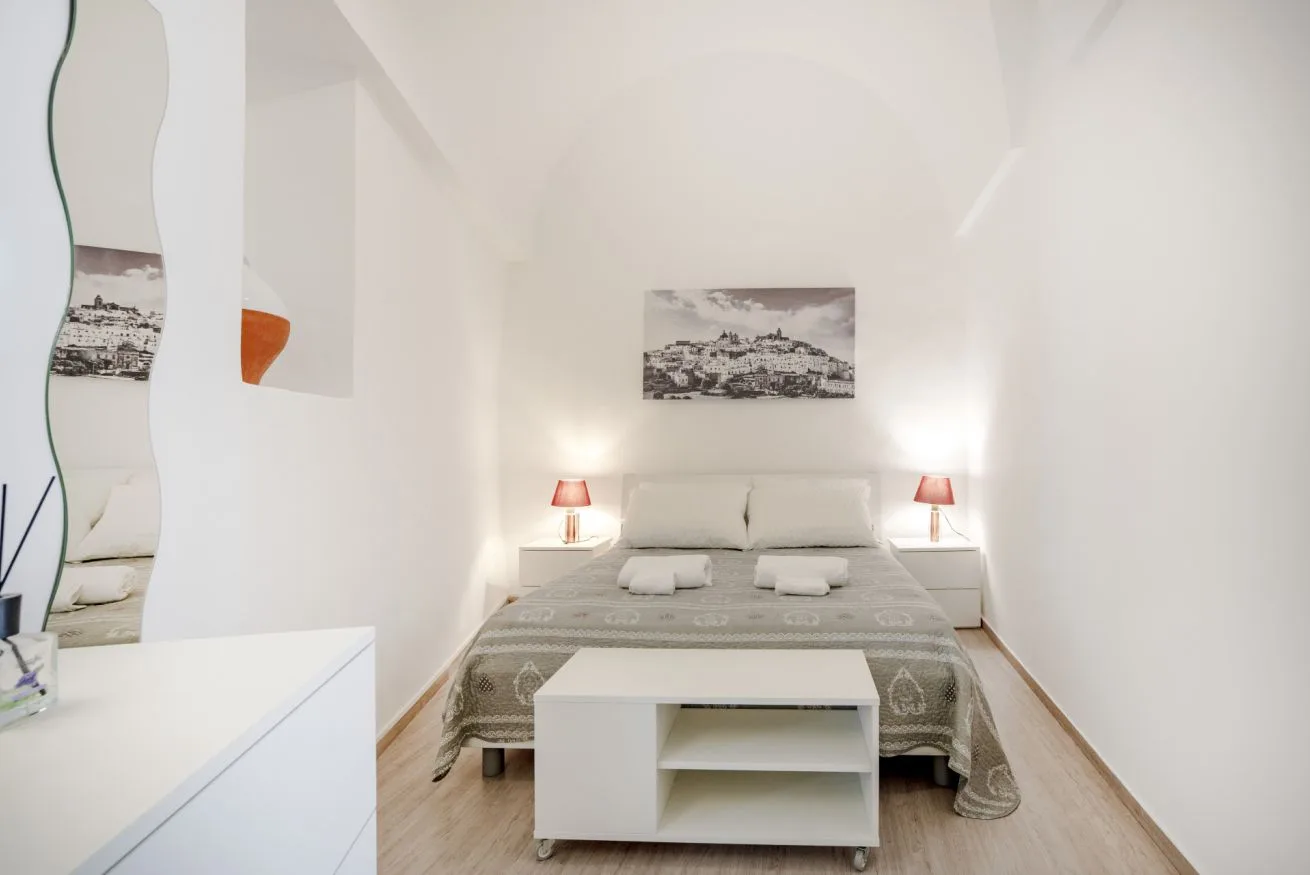 A bright bedroom with a double bed and a small table at its foot.