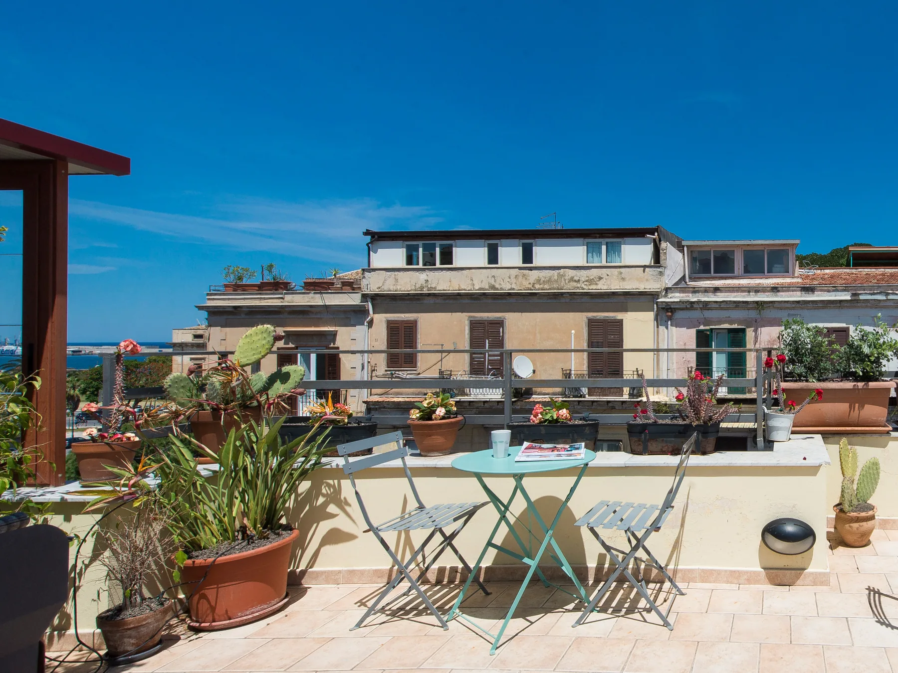 Una terraza soleada en la azotea tiene una mesa pequeña, sillas y plantas en macetas.