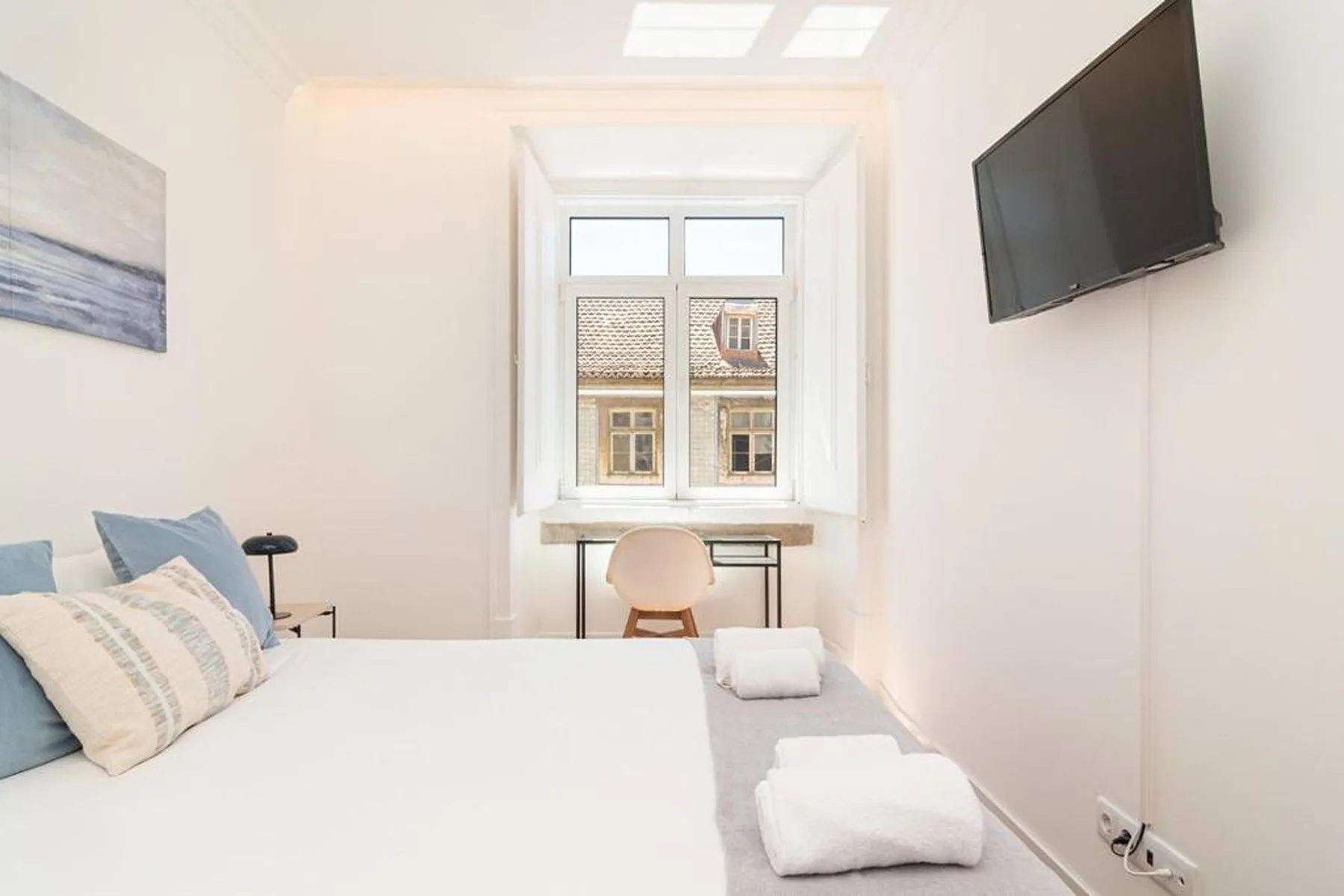 A bright bedroom with a neatly made bed, folded towels, and a window.