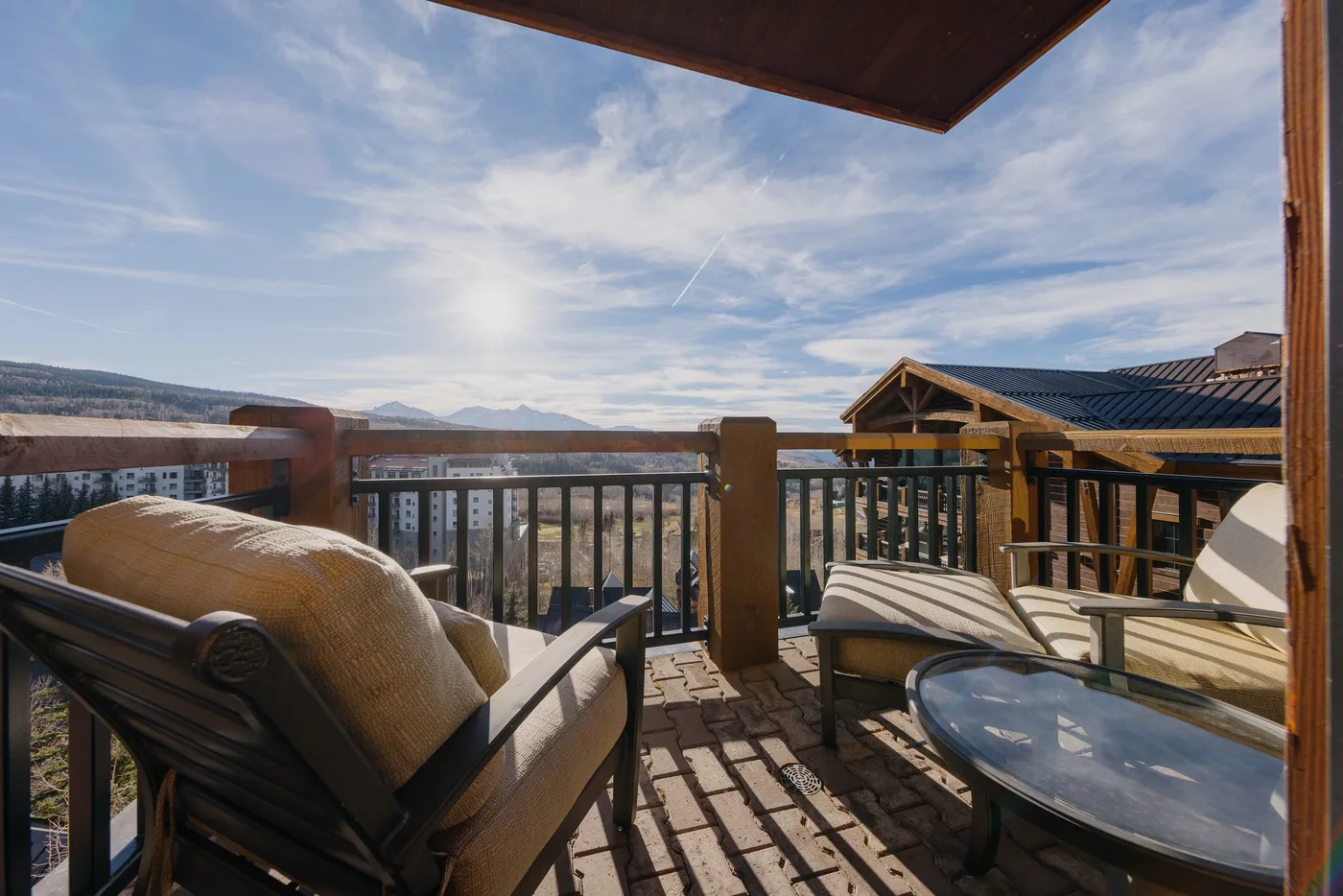 A sunlit outdoor deck with cushioned seating, a glass table, and a mountain view.