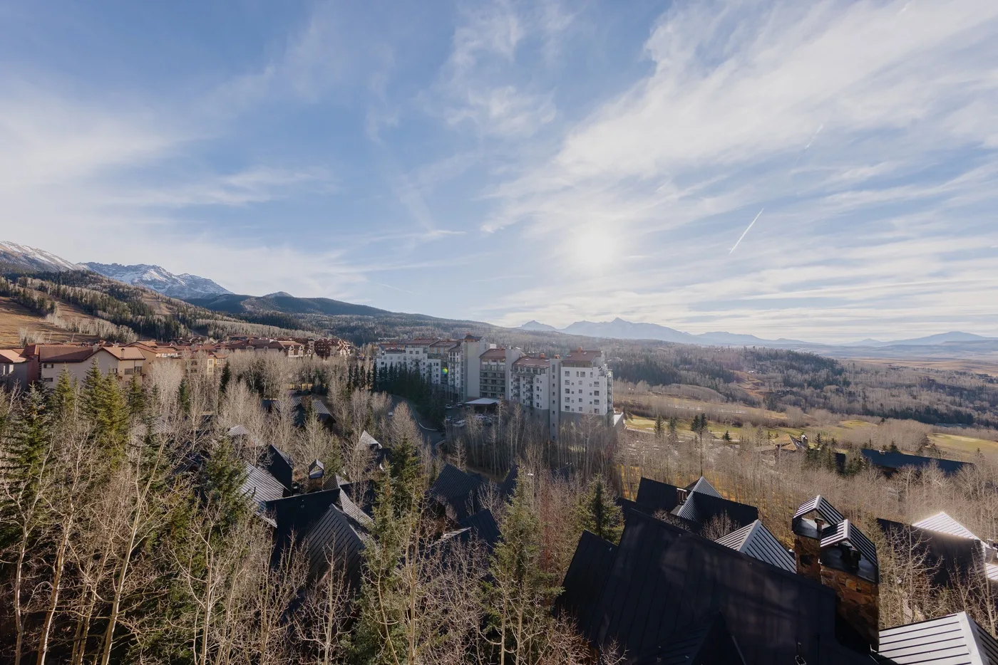A hillside town with apartment buildings, wooded slopes, and distant mountains beneath a broad sky.