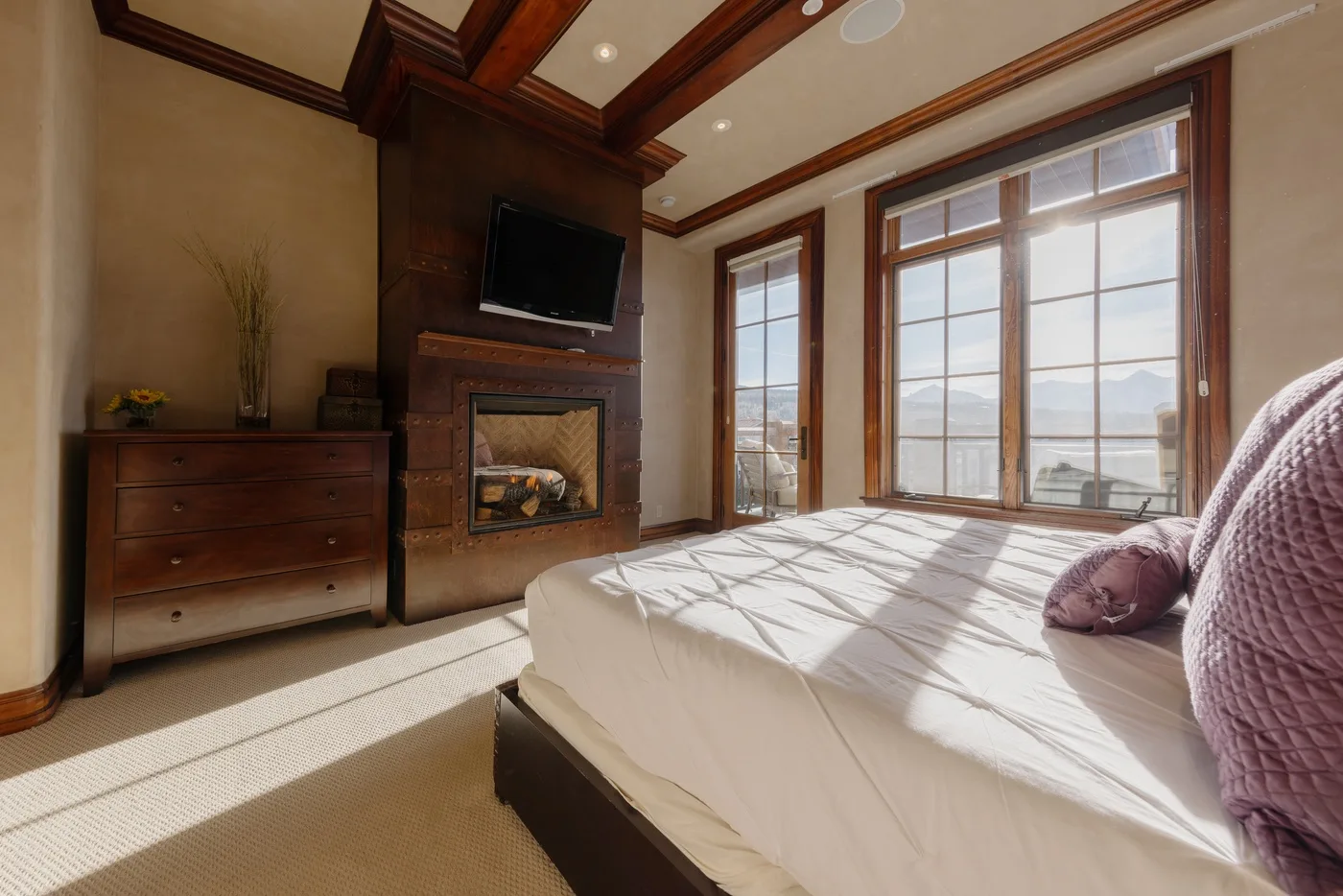 A sunlit bedroom with a bed beside a fireplace and large windows.