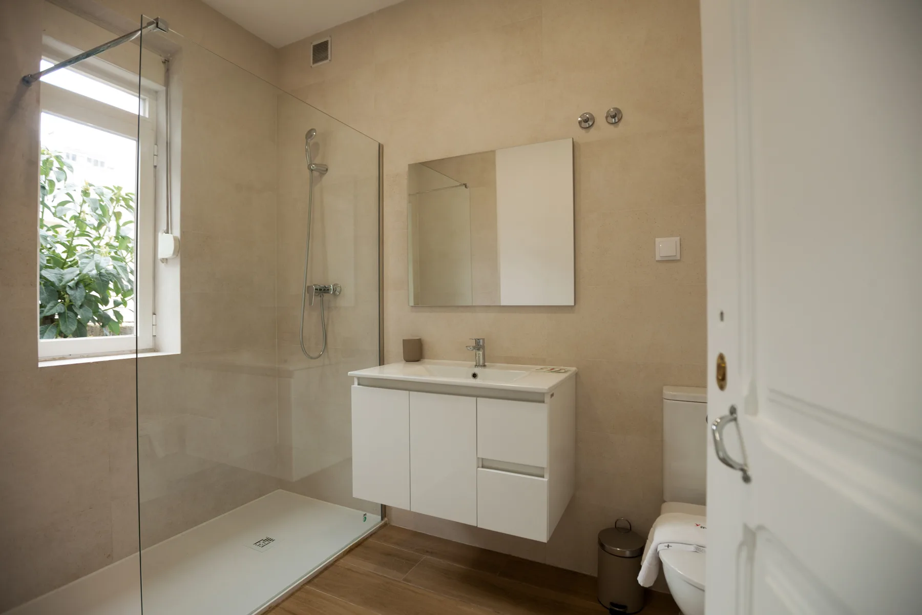 A bathroom with a glass shower, a vanity, and a window overlooking greenery.