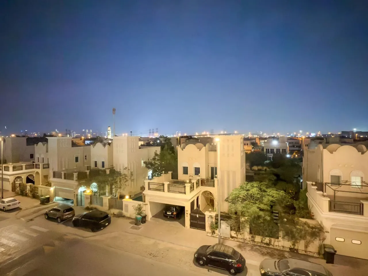 A nighttime streetscape of low-rise buildings, parked cars, and distant city lights.