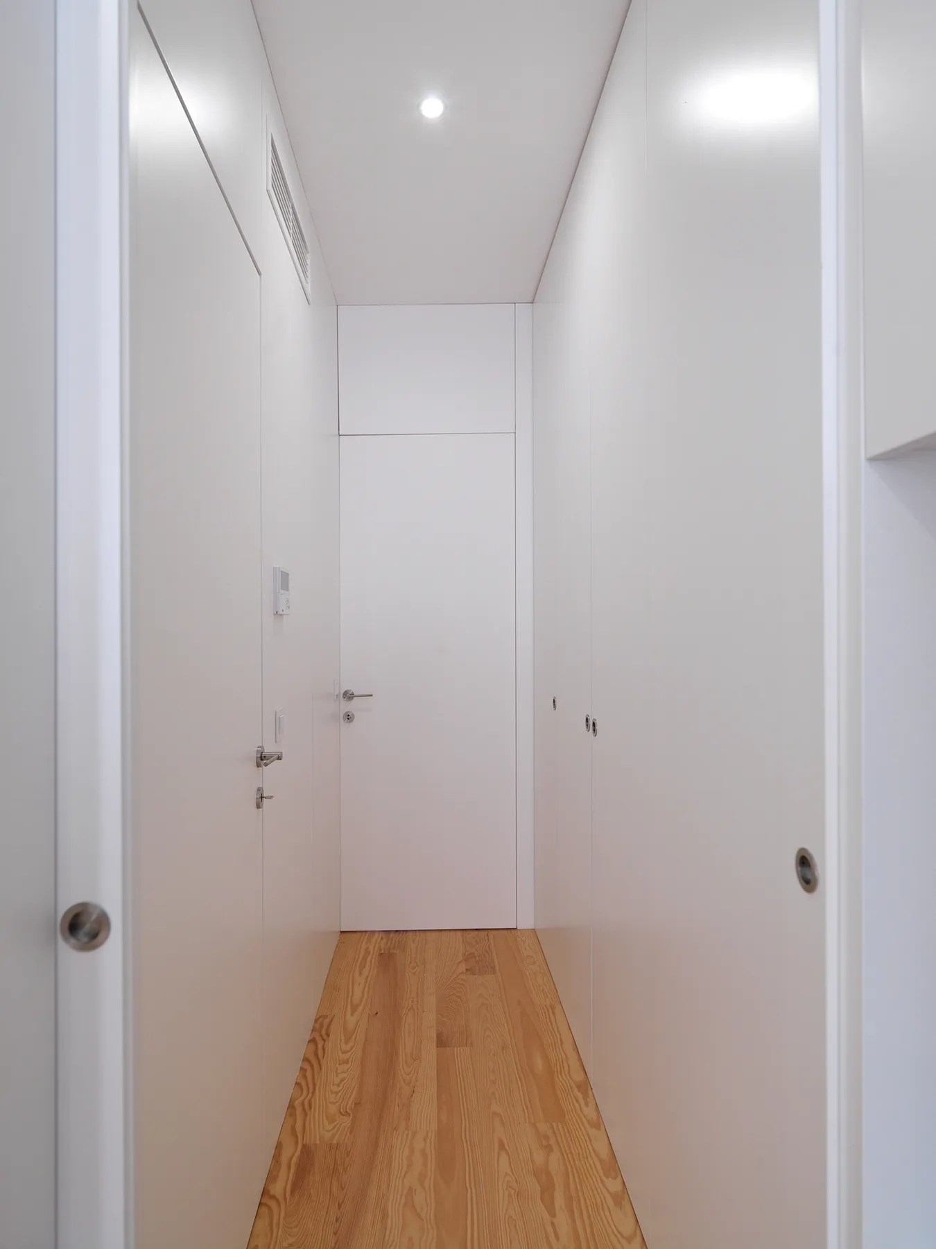 Un pasillo estrecho y blanco con suelo de madera clara y puertas cerradas.