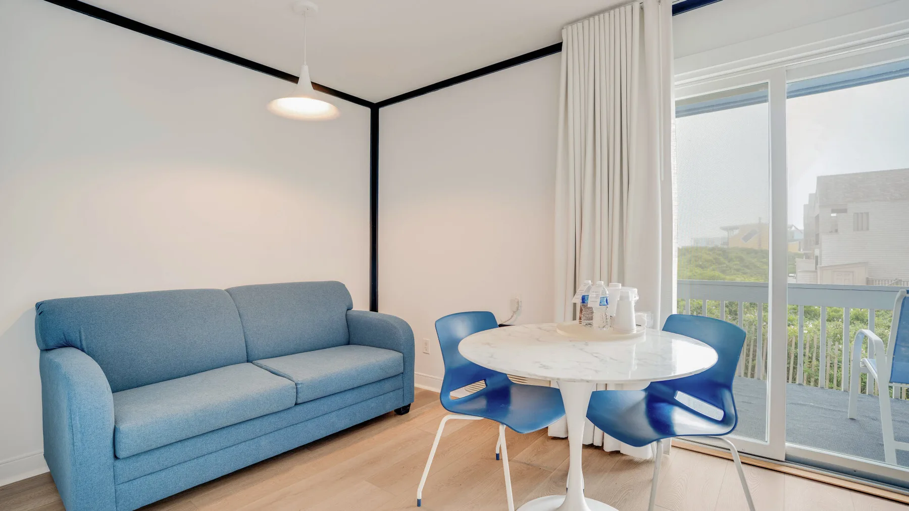 Una sala luminosa con asientos azules, una mesa redonda y una puerta de vidrio hacia el balcón.