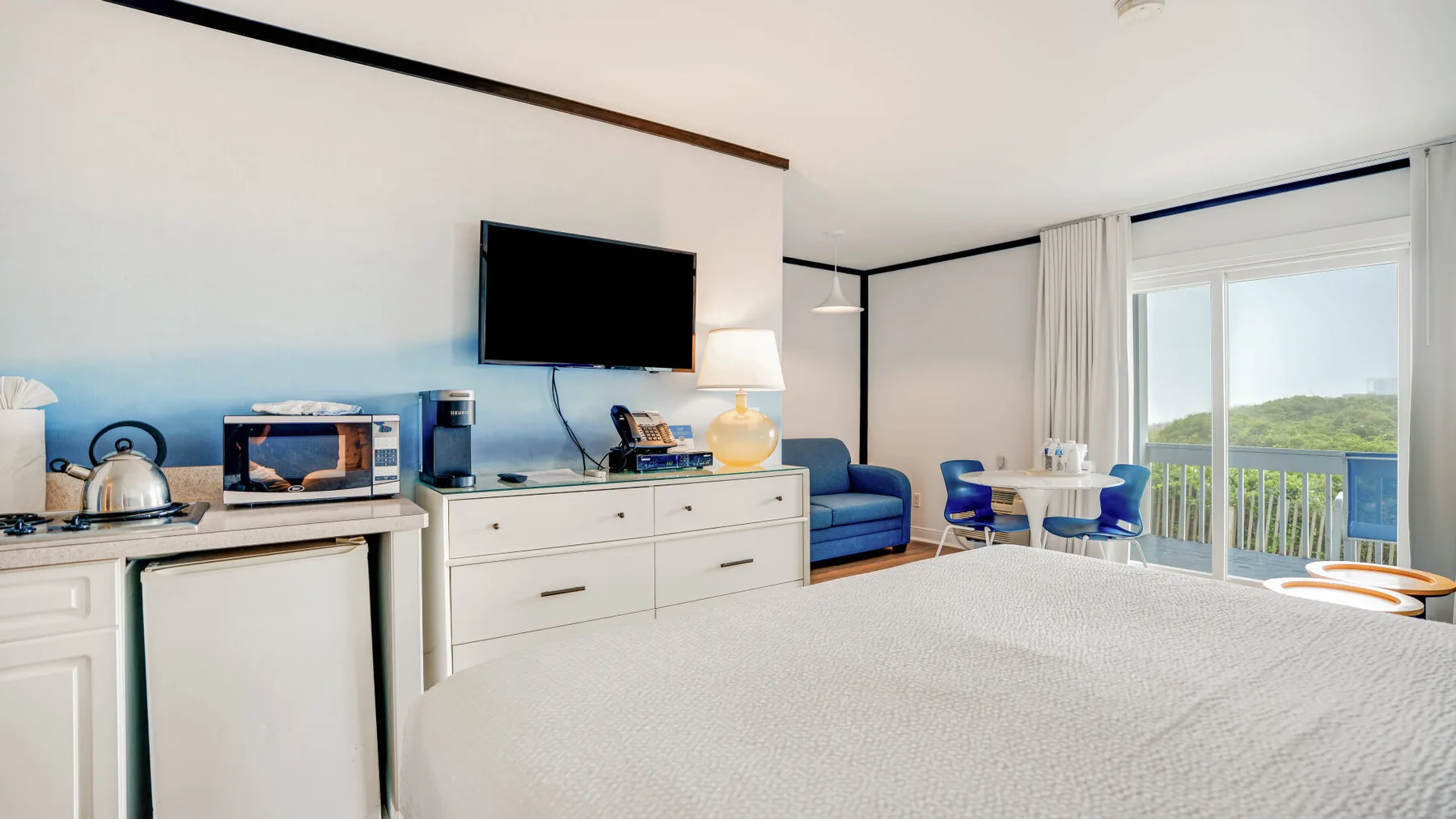 Una habitación luminosa con cocina pequeña, zona de estar y puertas corredizas de vidrio hacia un balcón.