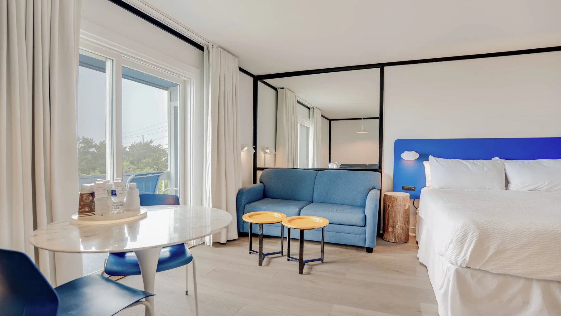 Un dormitorio luminoso con una zona de estar azul junto a la cama y grandes ventanas.