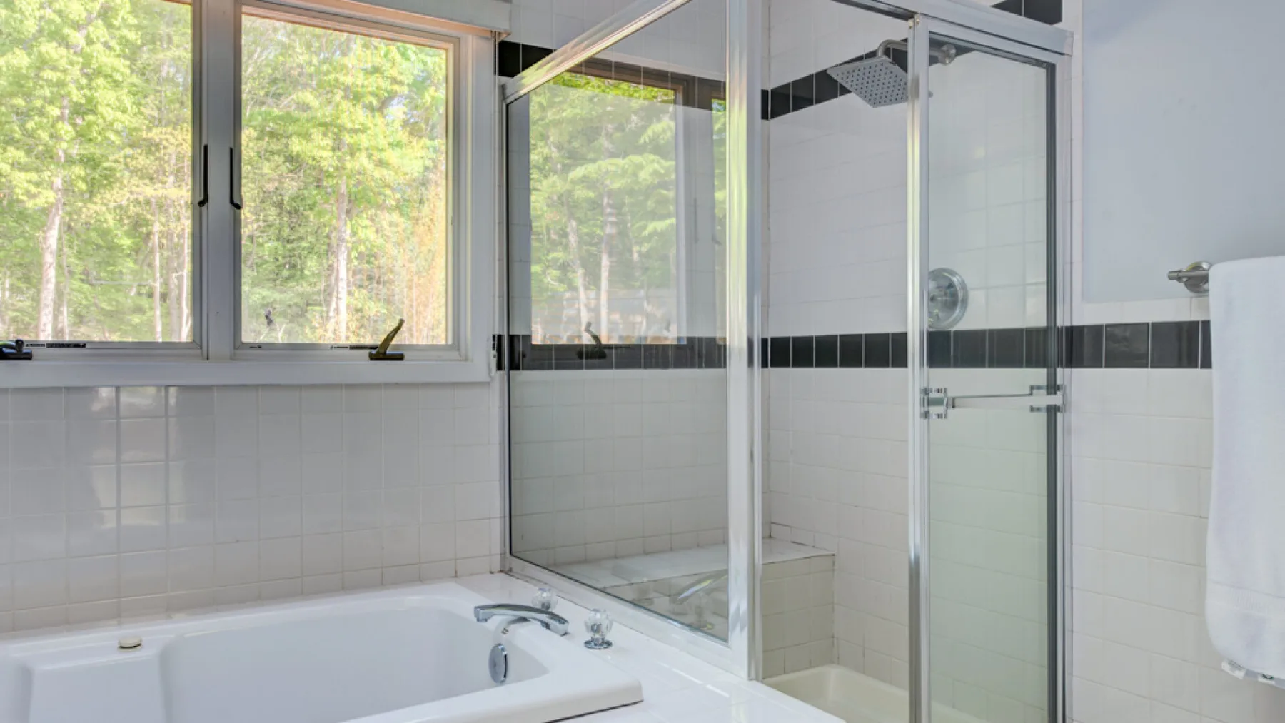 A bright bathroom with a bathtub, glass shower, and windows overlooking greenery.