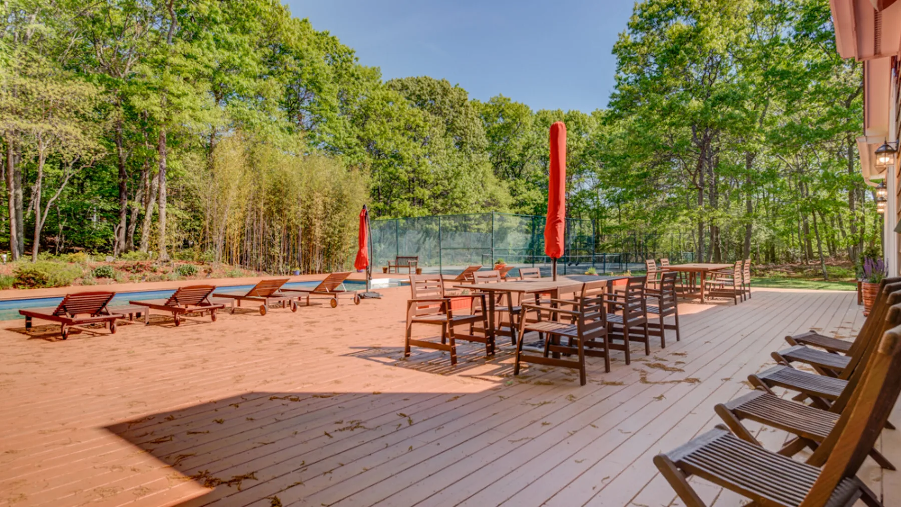 An outdoor wooden deck has seating and tables beside a fenced tennis court.