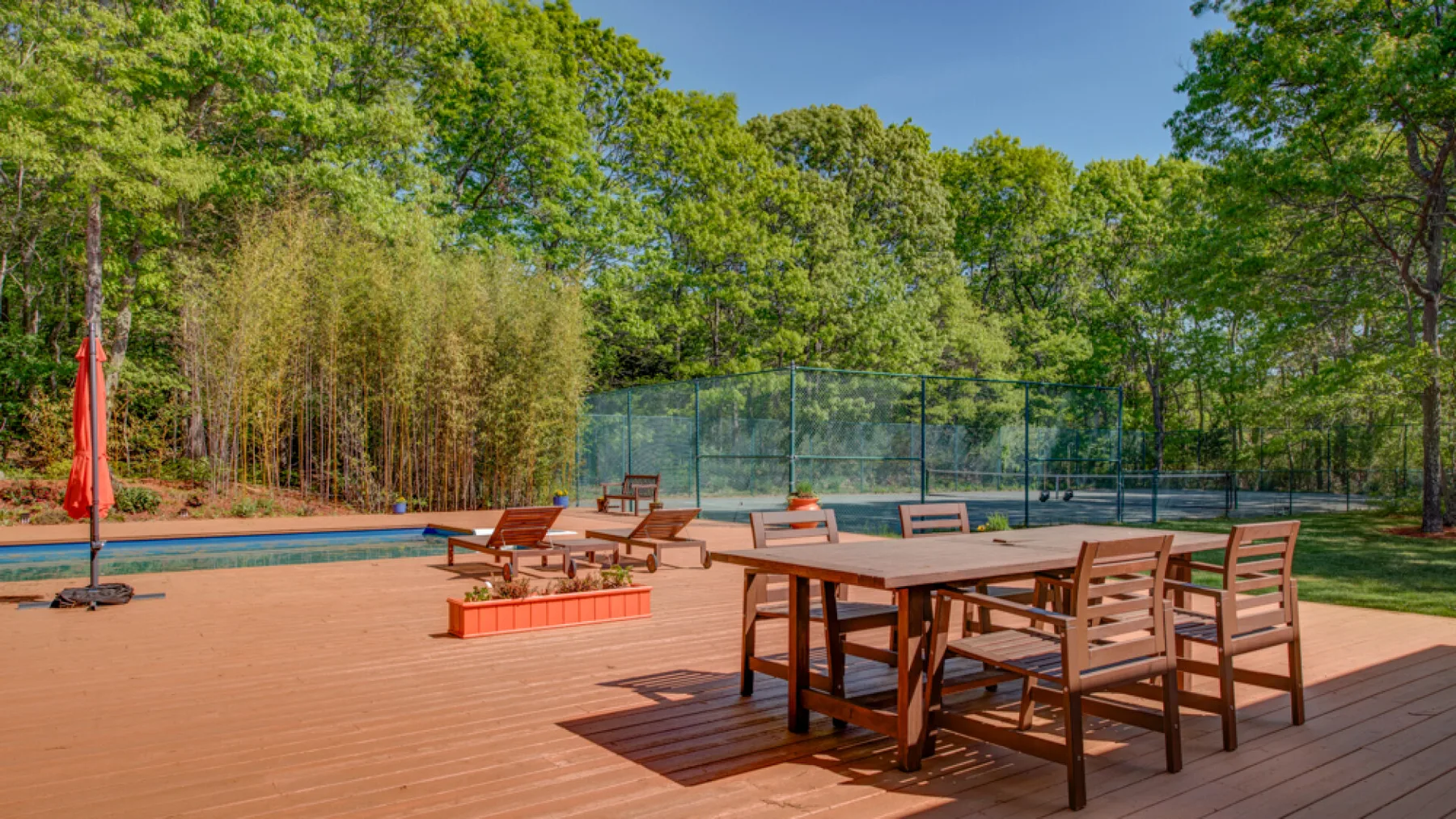 An outdoor patio with wooden seating and a fenced tennis court surrounded by trees.