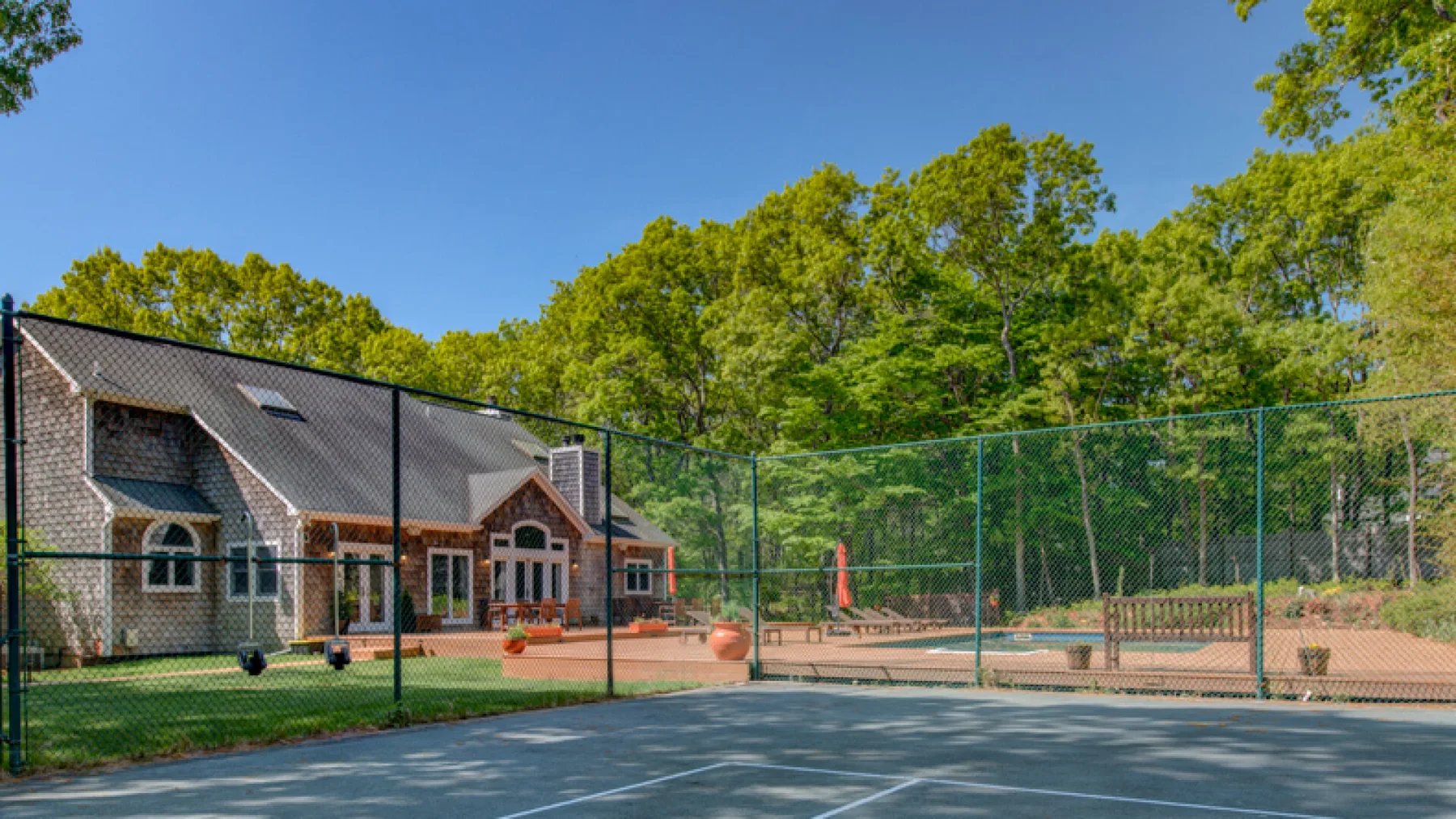 A fenced outdoor tennis court sits beside a brick building and surrounding trees.