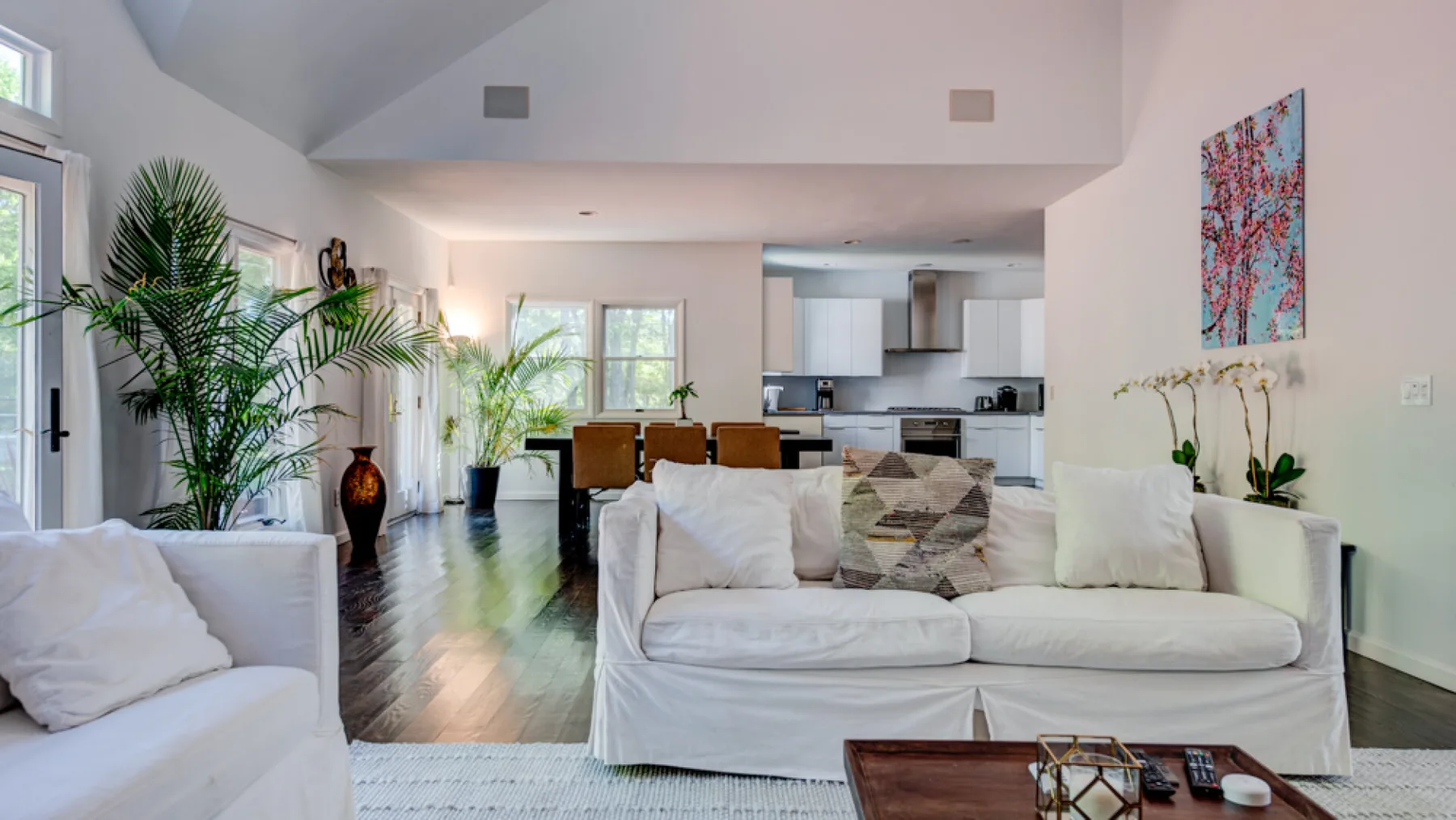 A bright open-plan living room with white seating and a kitchen in the background.