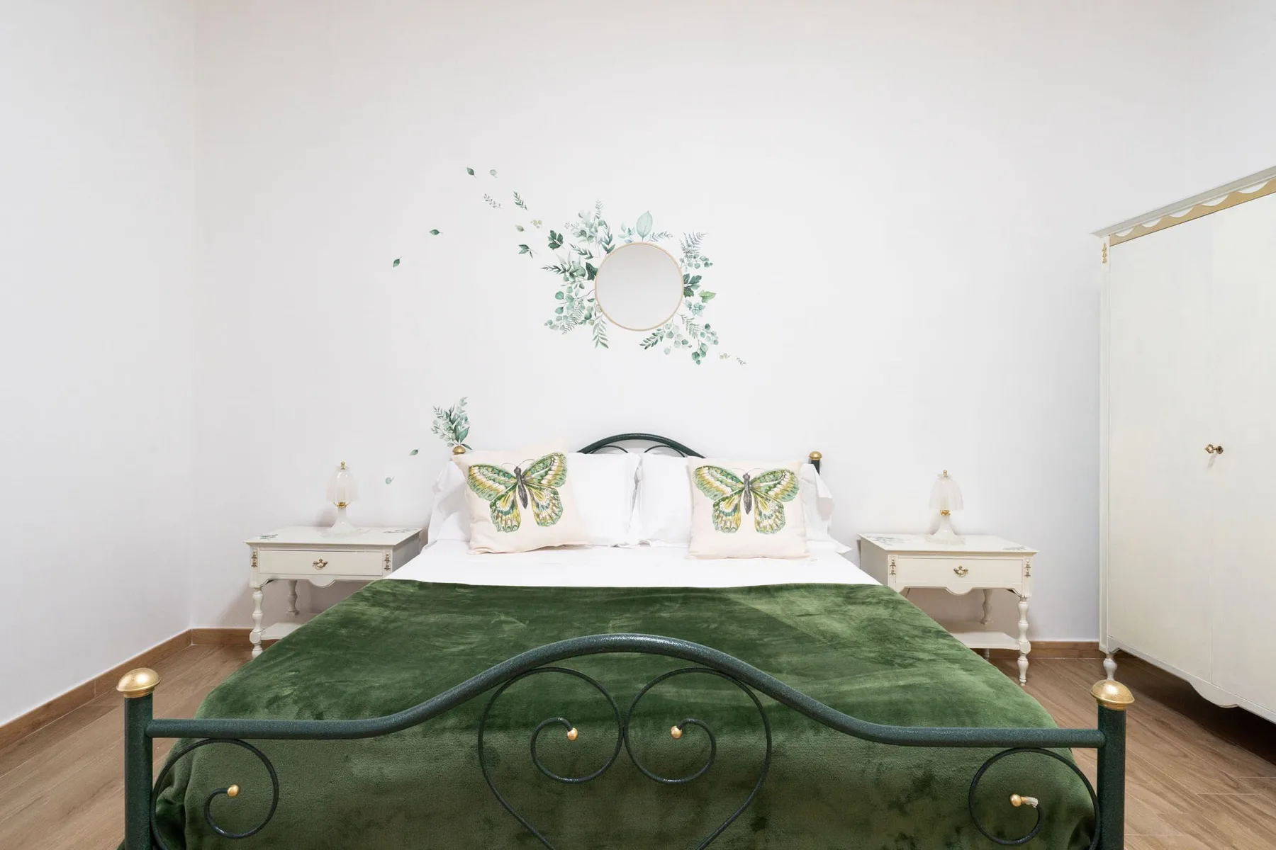 A bedroom with a green-covered bed, bedside tables, and a round wall mirror.