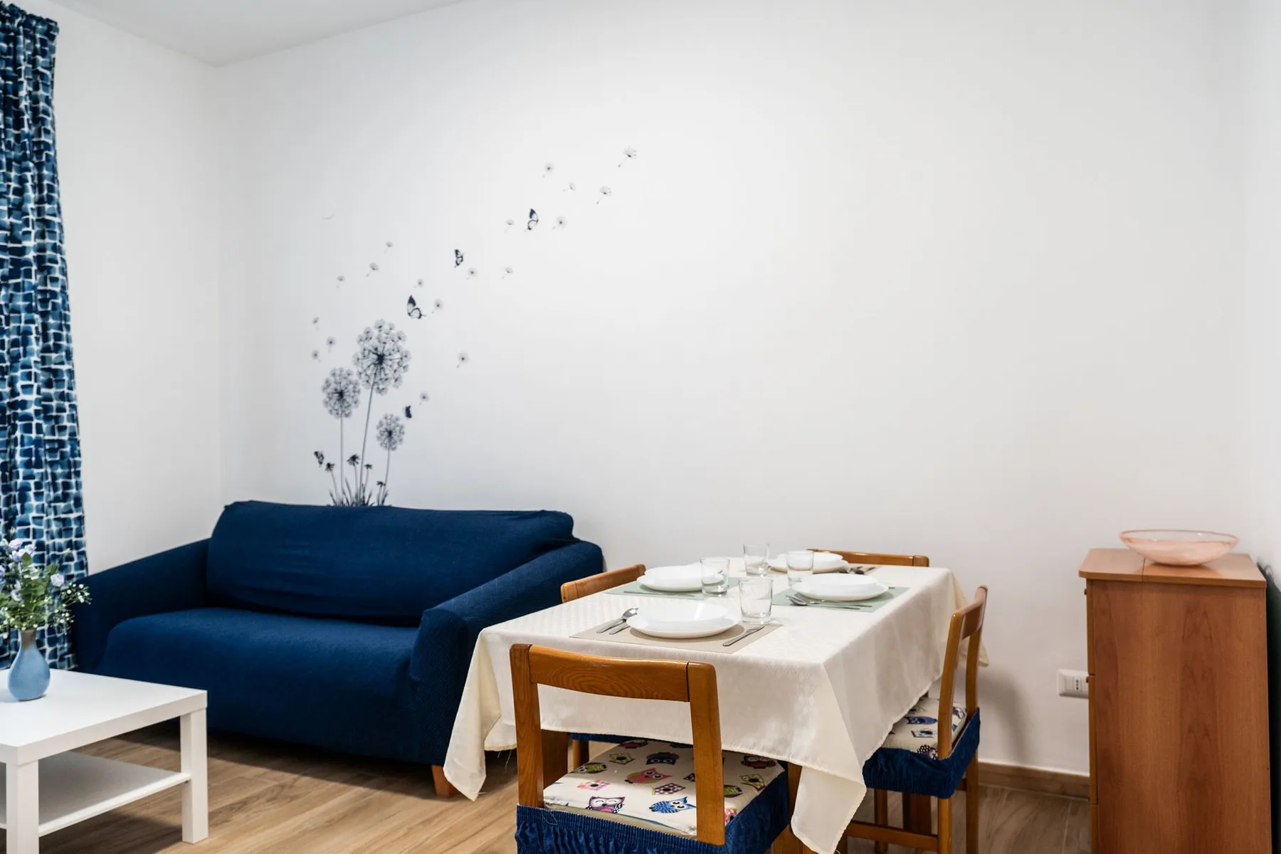 A dining table set with dishes stands beside blue seating in a bright room.