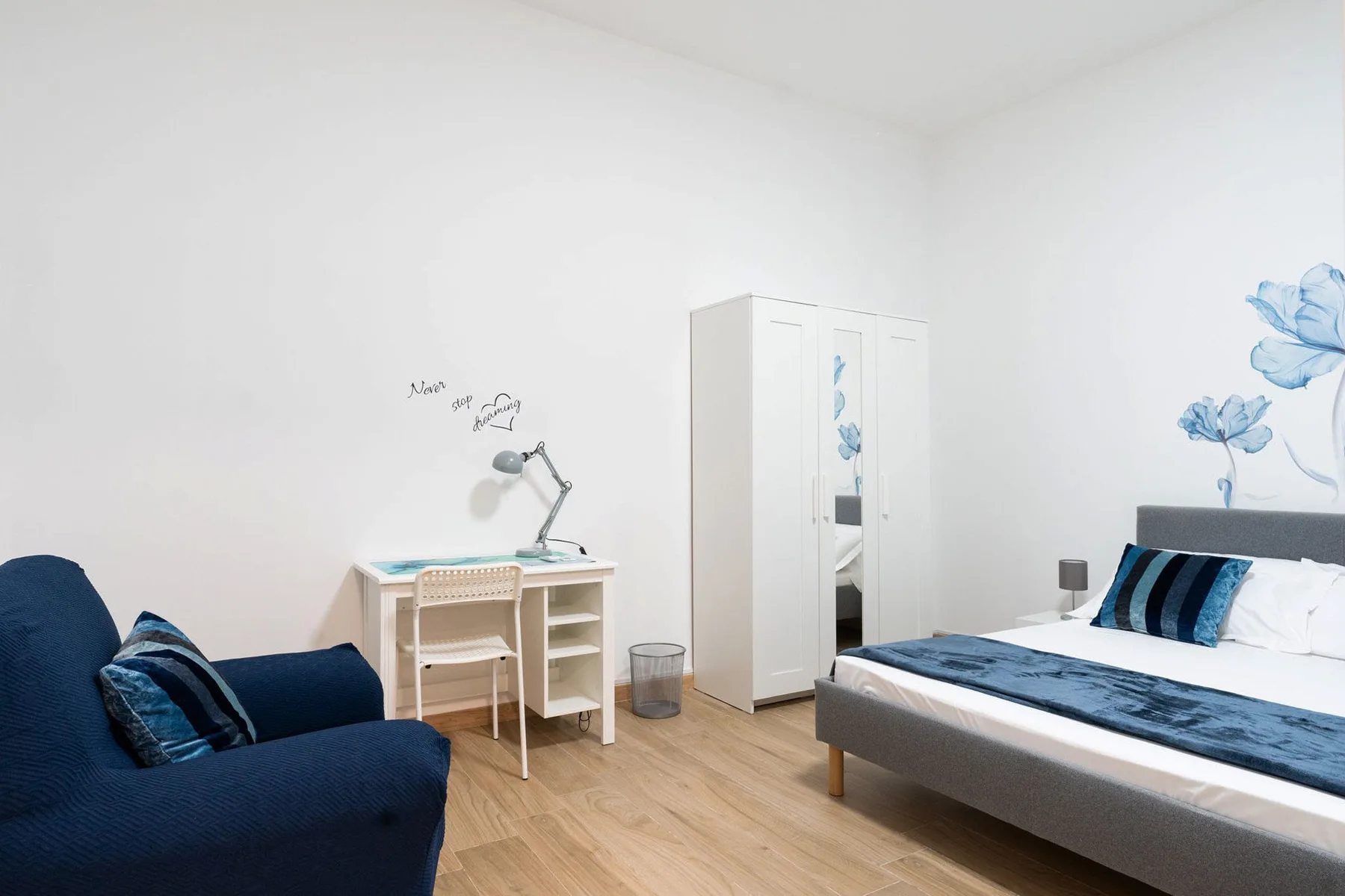 A bright bedroom with a bed, desk, and blue floral wall decoration.