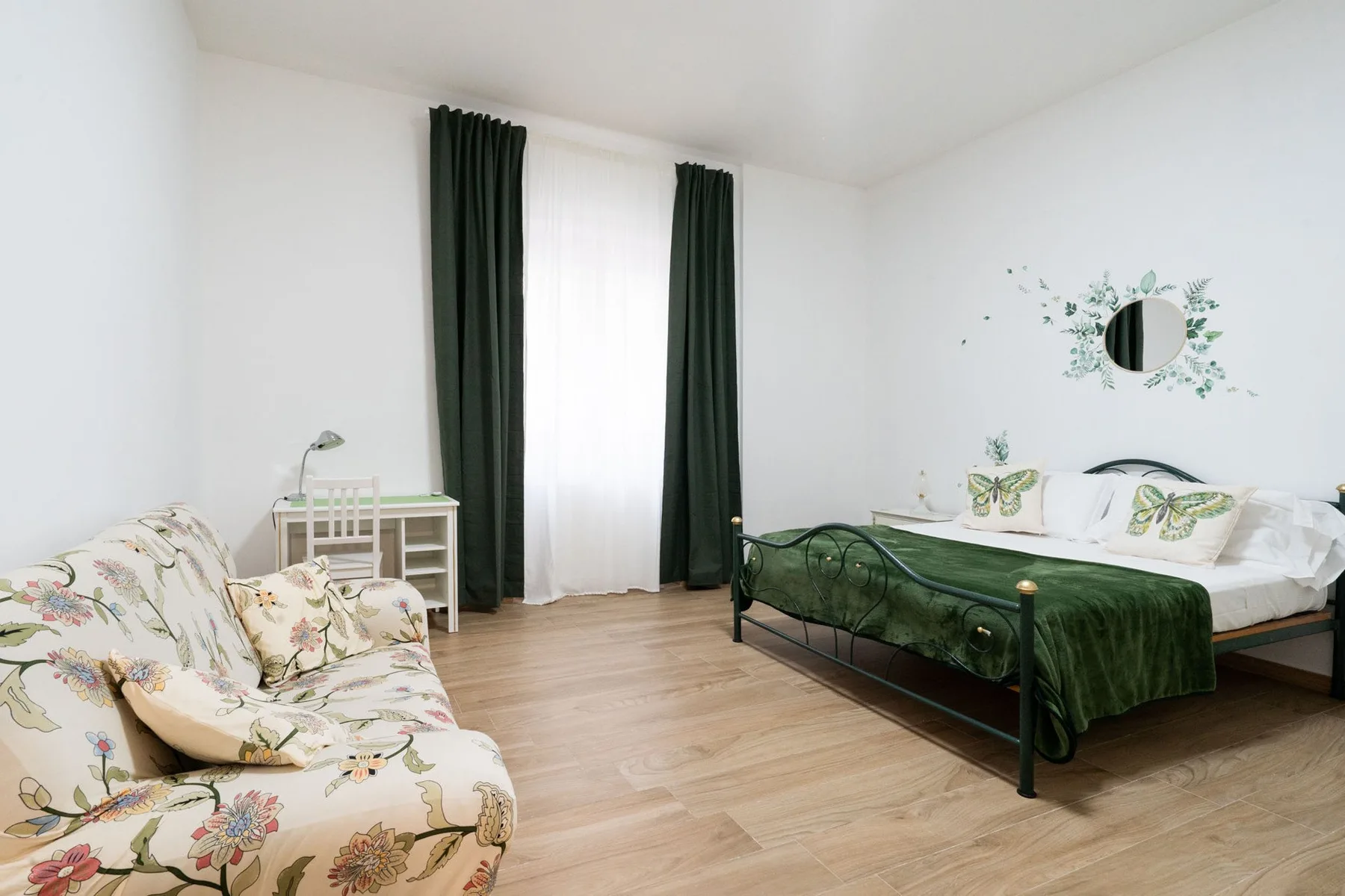 A bright bedroom with a bed, floral seating, and a tall curtained window.