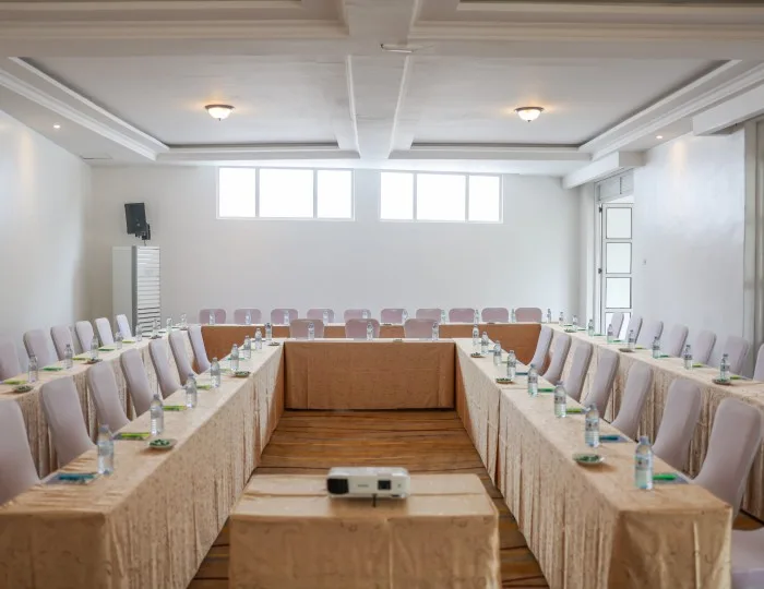 Una sala de conferencias con disposición en U, bajo ventanas que dejan entrar luz natural, equipada con blocs de notas, agua y un proyector.