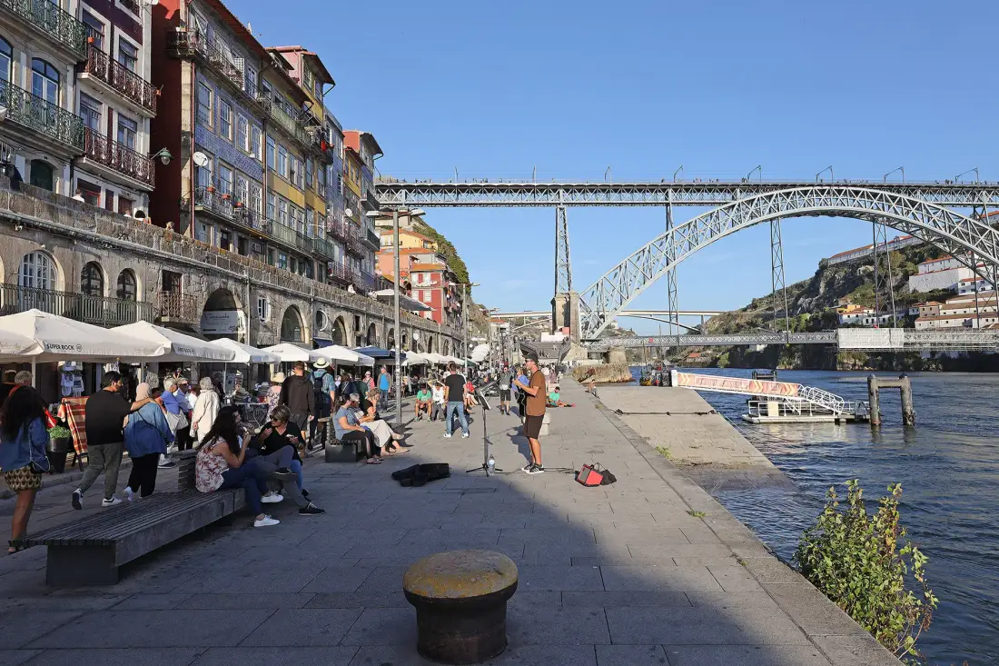 A riverside promenade with people walking and a large bridge in the background.