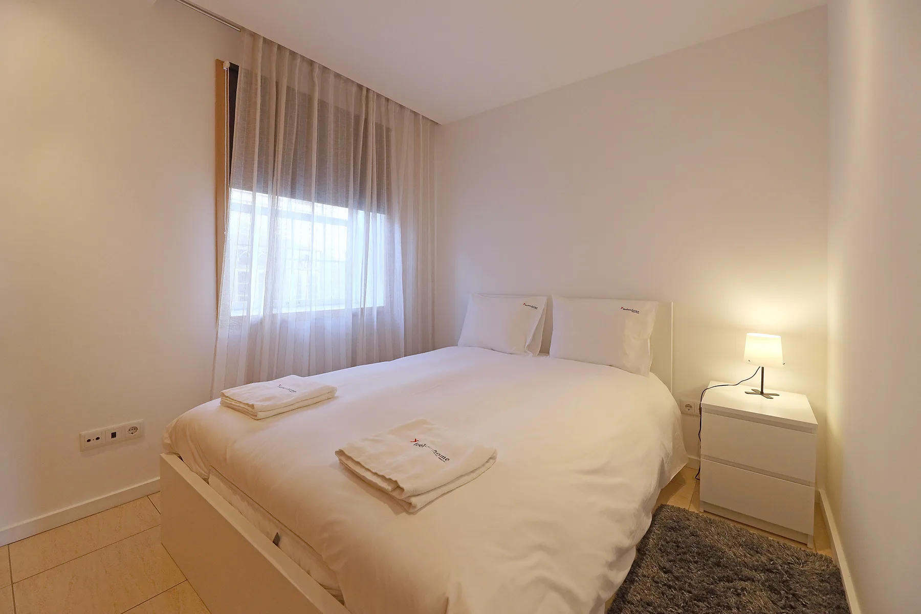 A bedroom with a neatly made bed and a window with sheer curtains.