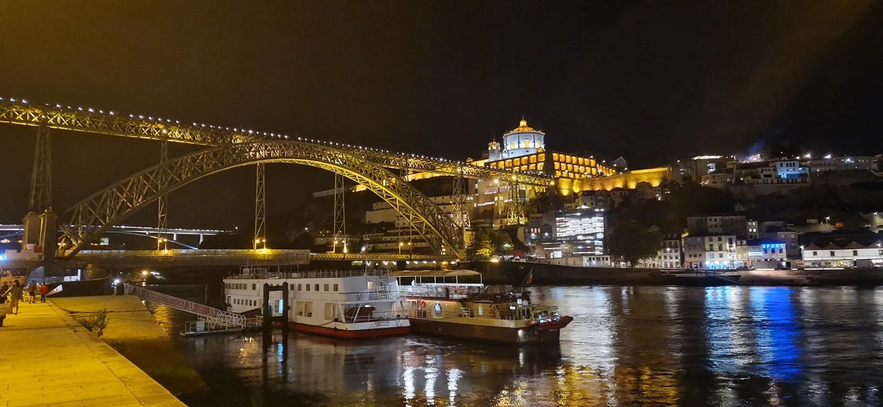 A nighttime riverfront scene shows a lit bridge, boats, and illuminated buildings along the water.