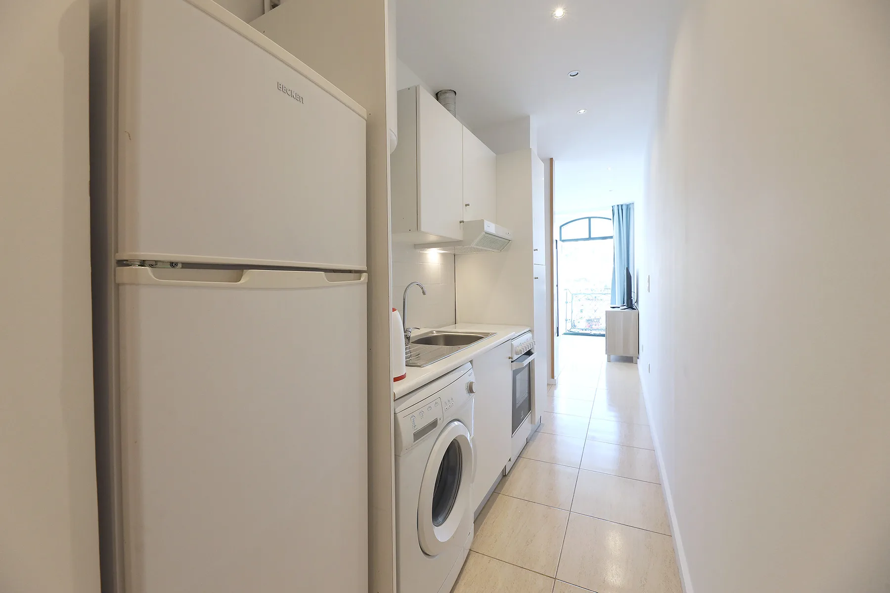 A narrow kitchen with white cabinets and appliances opens toward a bright window.