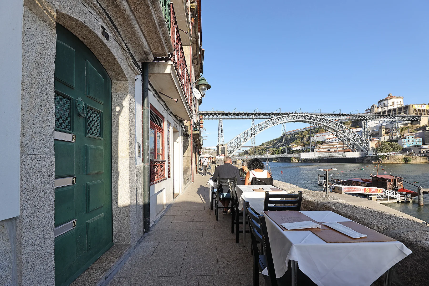 Outdoor dining tables line a riverside walkway beneath a large arched bridge.
