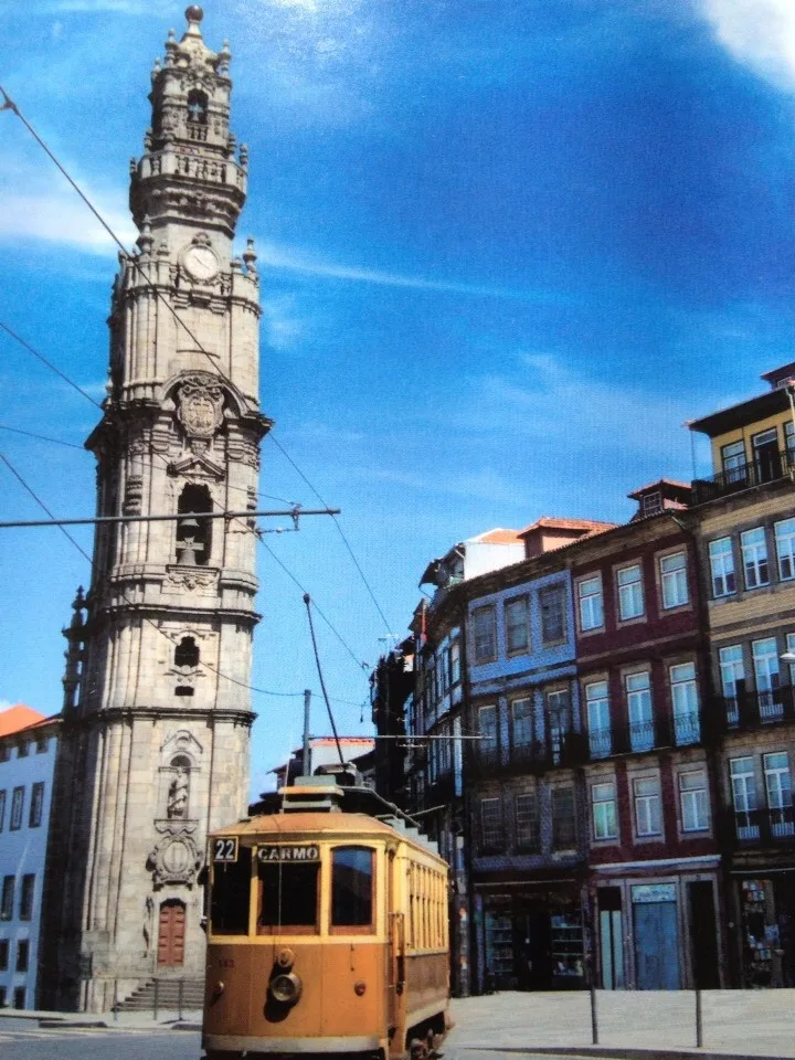 A stone clock tower rises above a city street as a yellow tram passes.