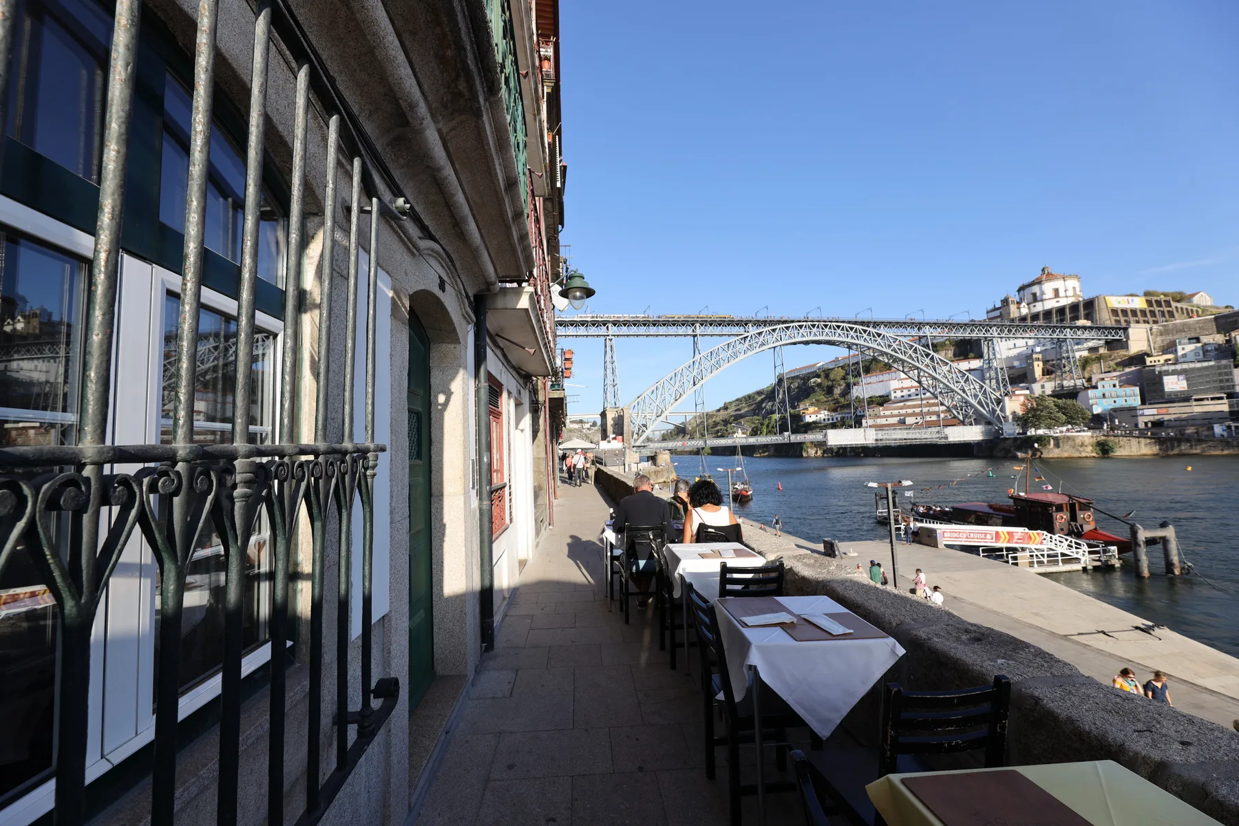 Riverside café terrace with outdoor tables overlooking a bridge and the water.