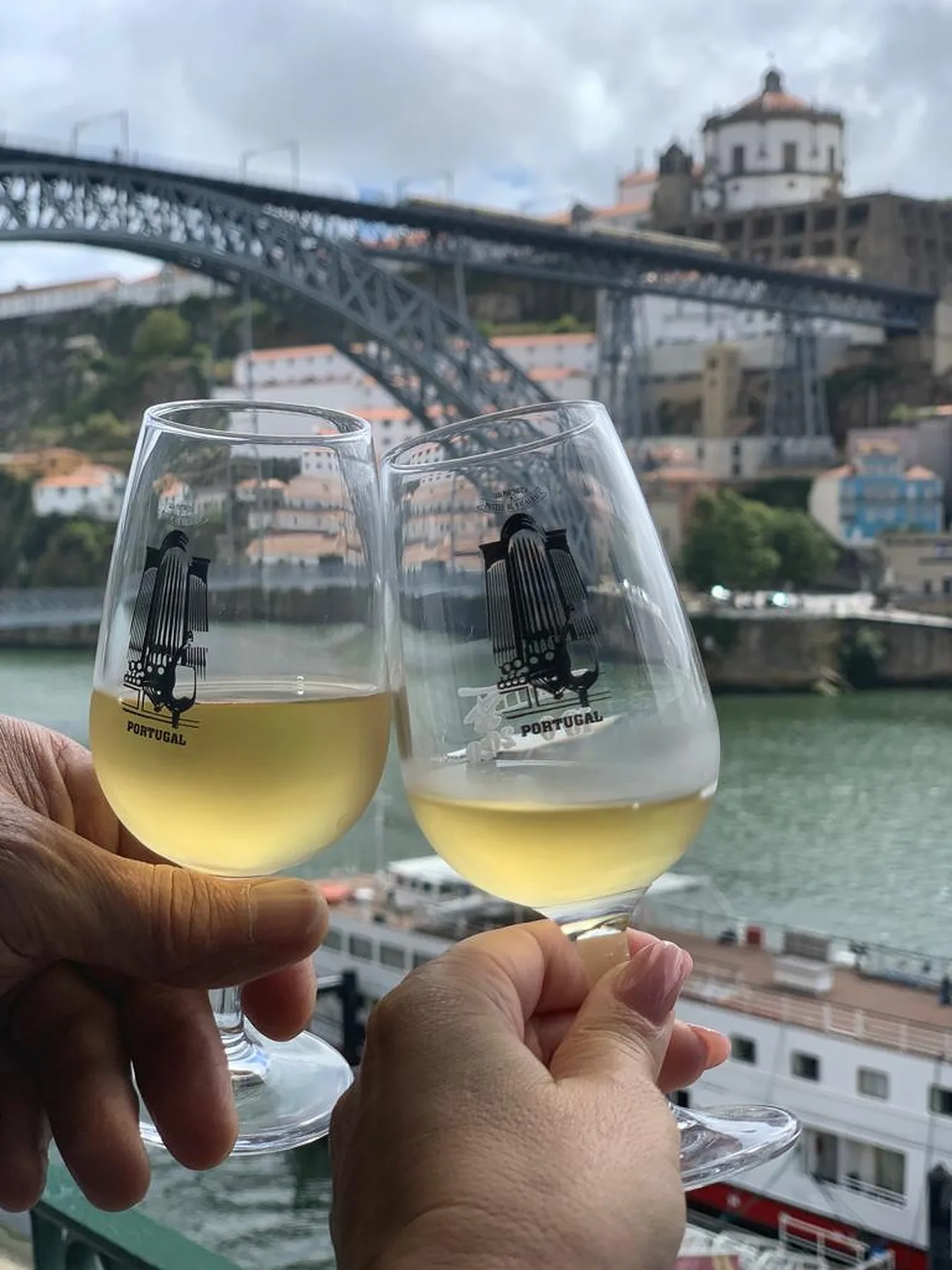 Two glasses of white wine are raised above a riverside view of a bridge and buildings.
