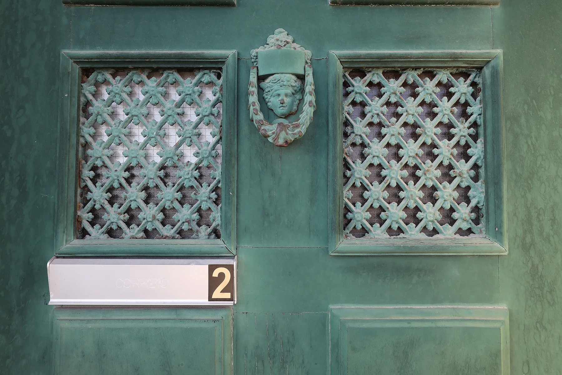 A weathered green door has two decorative lattice panels and a sculpted face.