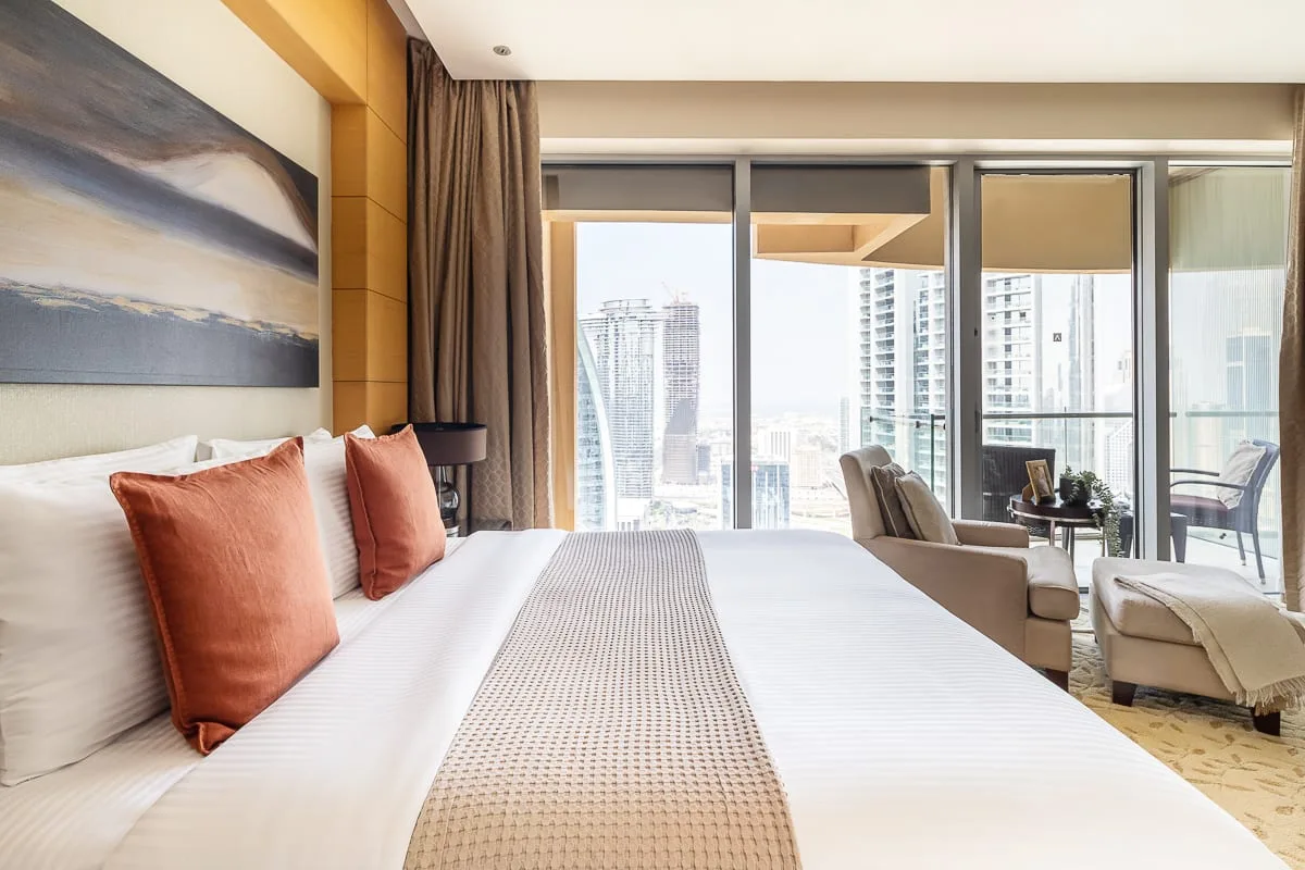 Un dormitorio con una cama bien tendida y grandes ventanas con vistas a edificios de la ciudad.