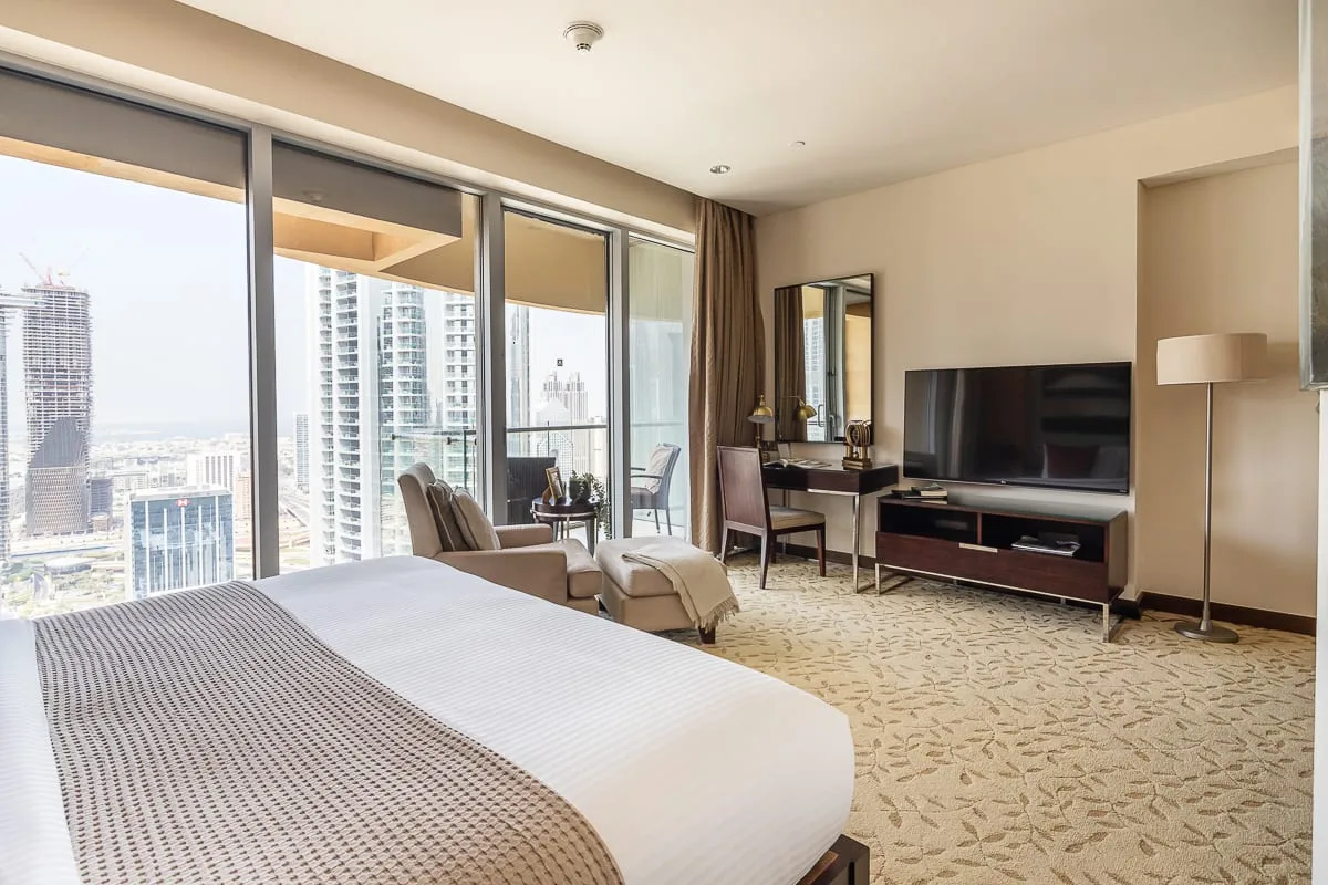 Un dormitorio con una cama grande, una zona de estar y ventanales con vistas a edificios de la ciudad.