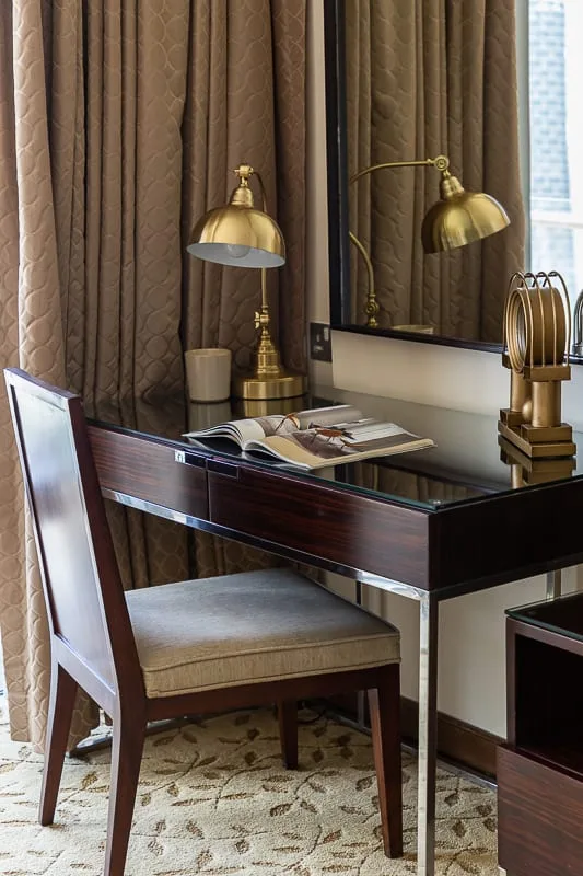 Un escritorio y una silla junto a ventanas con cortinas, con una lámpara de latón sobre el escritorio.