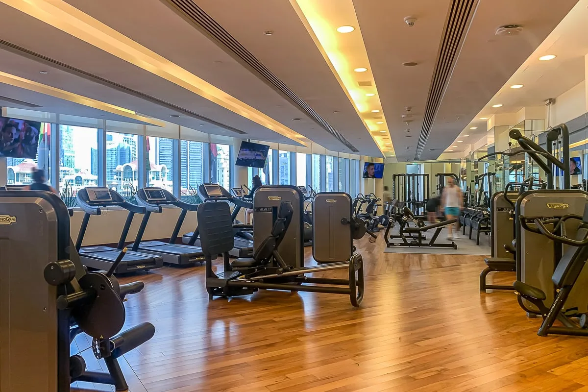 Un gimnasio con máquinas de ejercicio y cintas de correr junto a grandes ventanas.