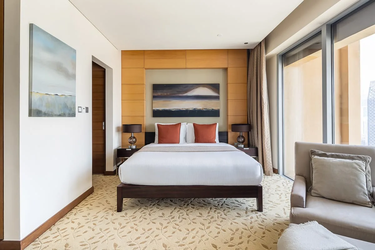 Un dormitorio con una cama bien tendida, una obra enmarcada y grandes ventanas.