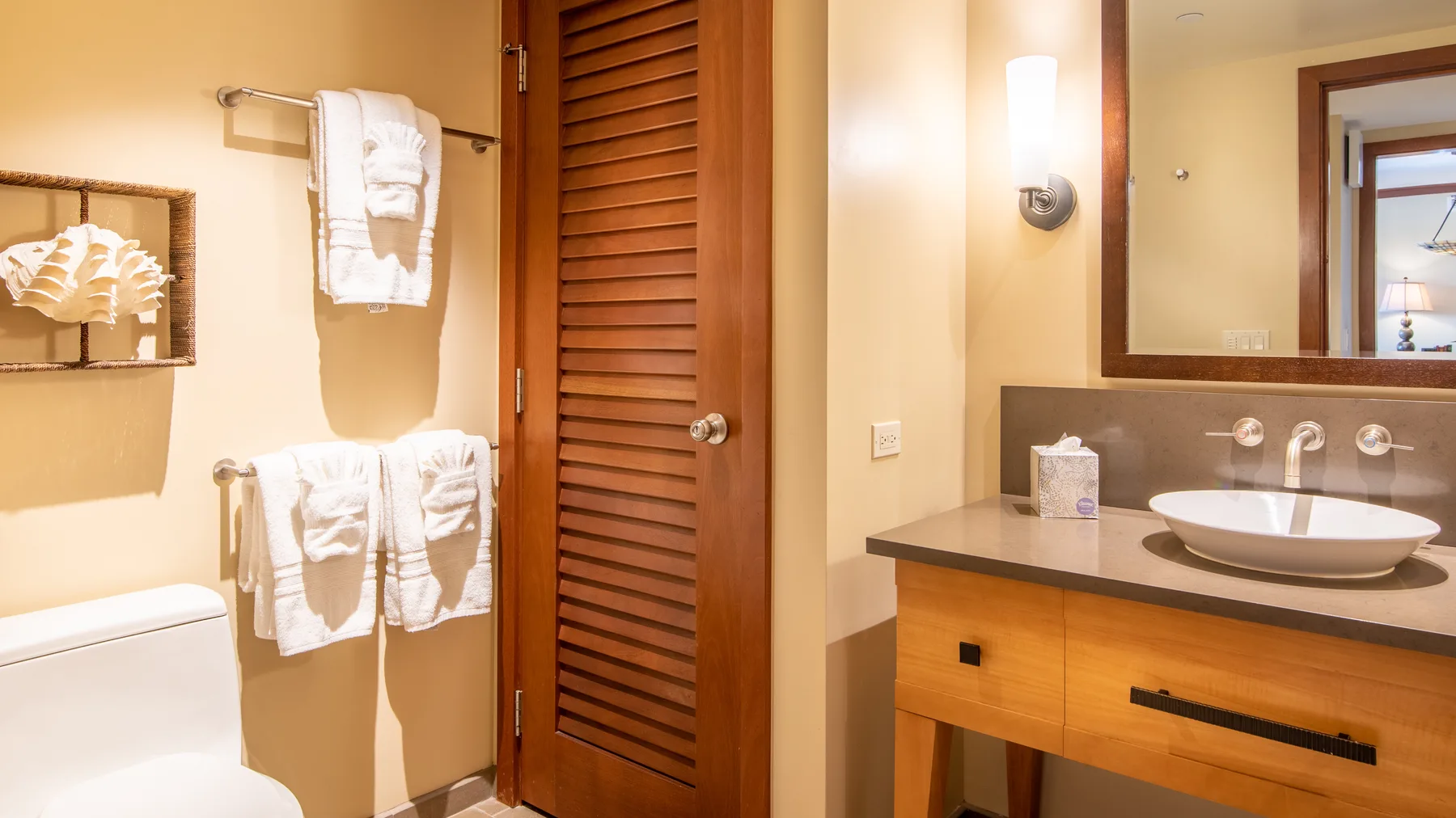 Un tocador de baño con lavabo sobre encimera, espejo y puerta de madera con lamas.