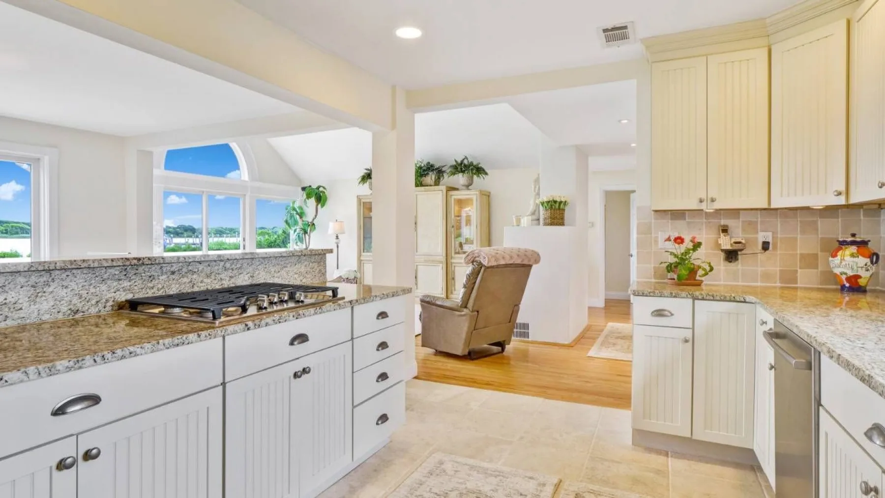 A bright kitchen with white cabinetry and a central island opens onto a living area.