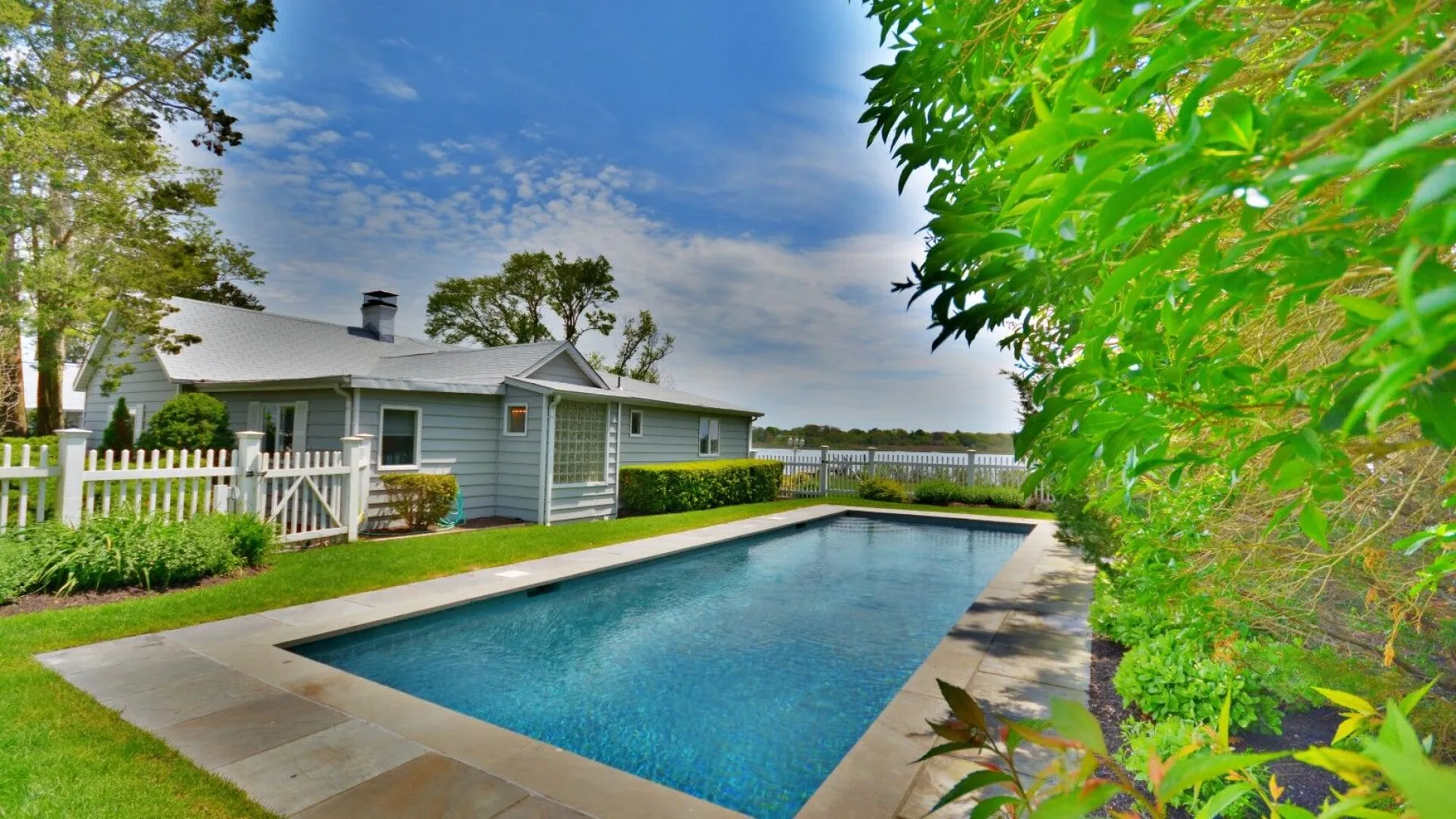 An outdoor swimming pool beside a low house, with leafy trees nearby.