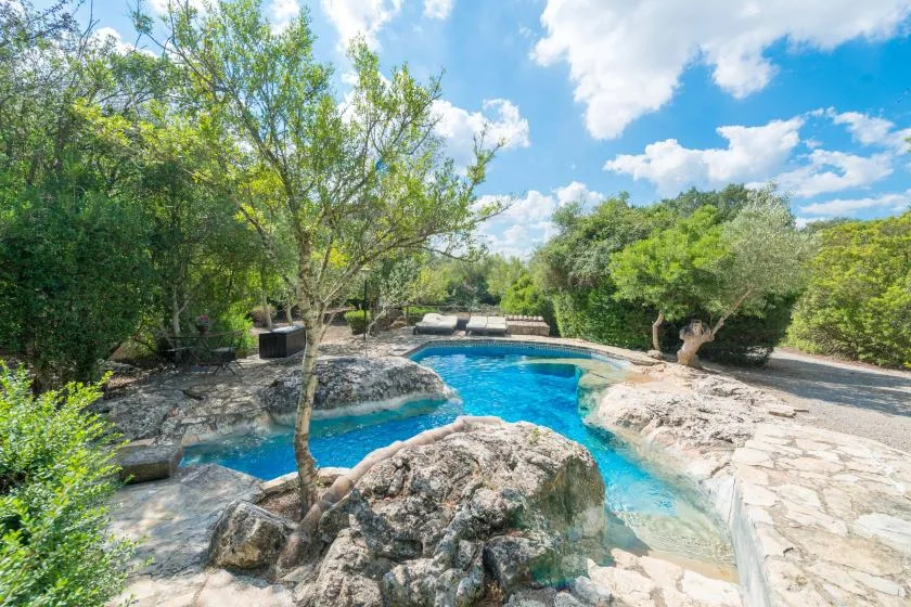 Una piscina al aire libre está rodeada de rocas y árboles bajo un cielo despejado.