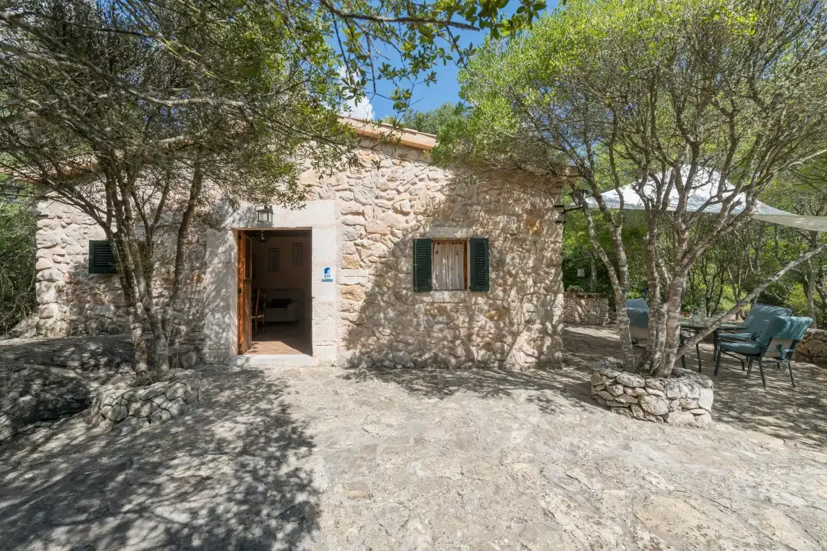 La entrada de una casa de piedra da a un patio exterior sombreado con árboles y asientos.