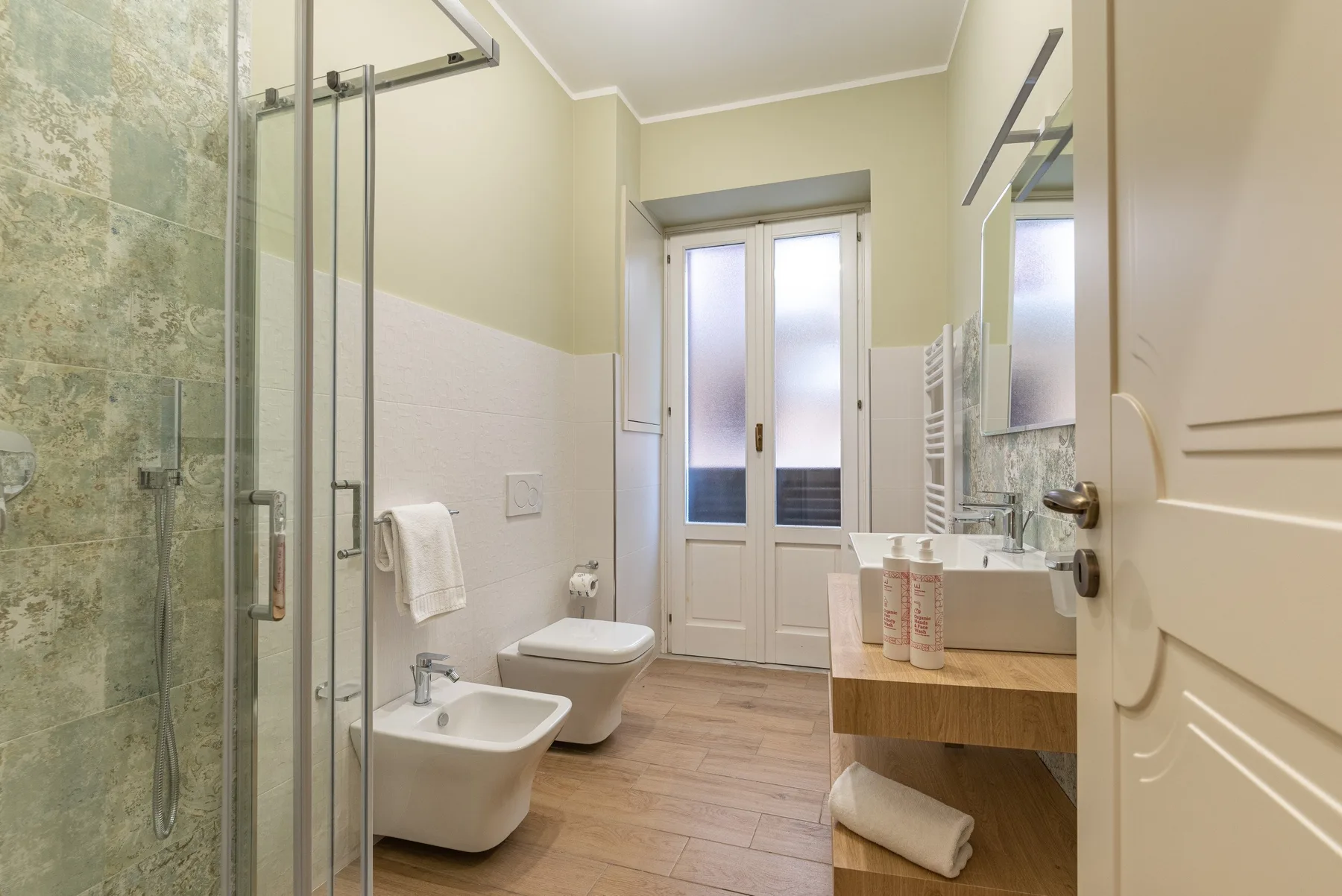 Un baño luminoso con ducha de vidrio, lavabo, inodoro y puertas a un balcón.