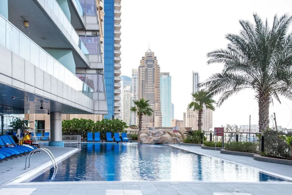 Una piscina al aire libre junto a un edificio moderno, con palmeras y torres urbanas al fondo.