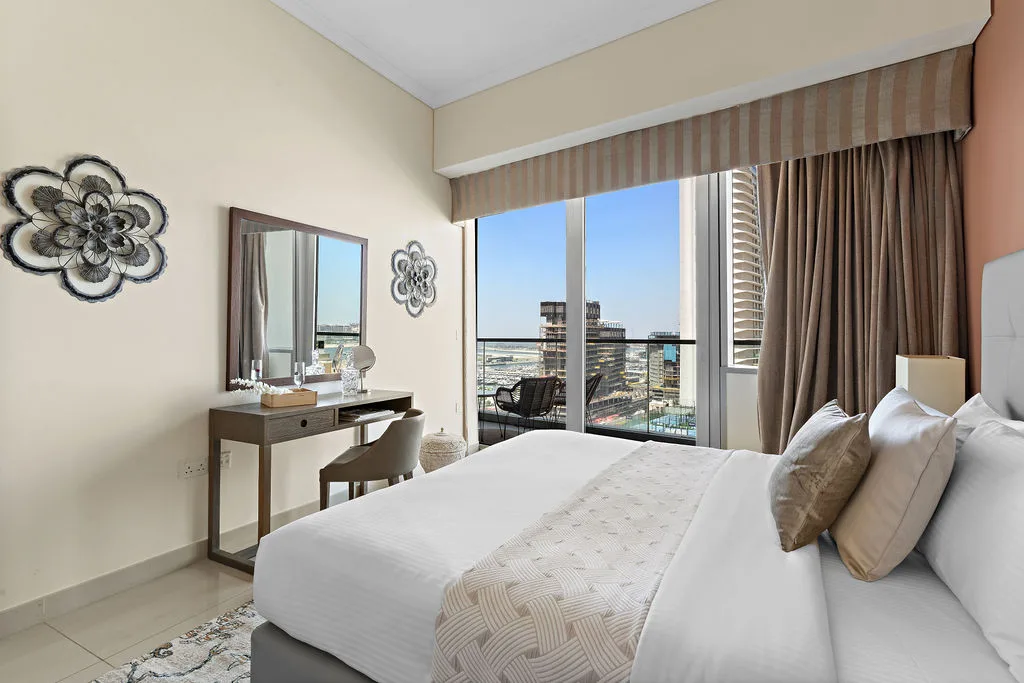 Un dormitorio luminoso con una cama tendida y ventanas de piso a techo con vistas a la ciudad.