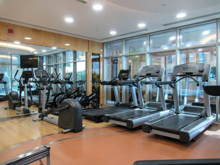 Un gimnasio con cintas de correr y otras máquinas de ejercicio junto a grandes ventanales.